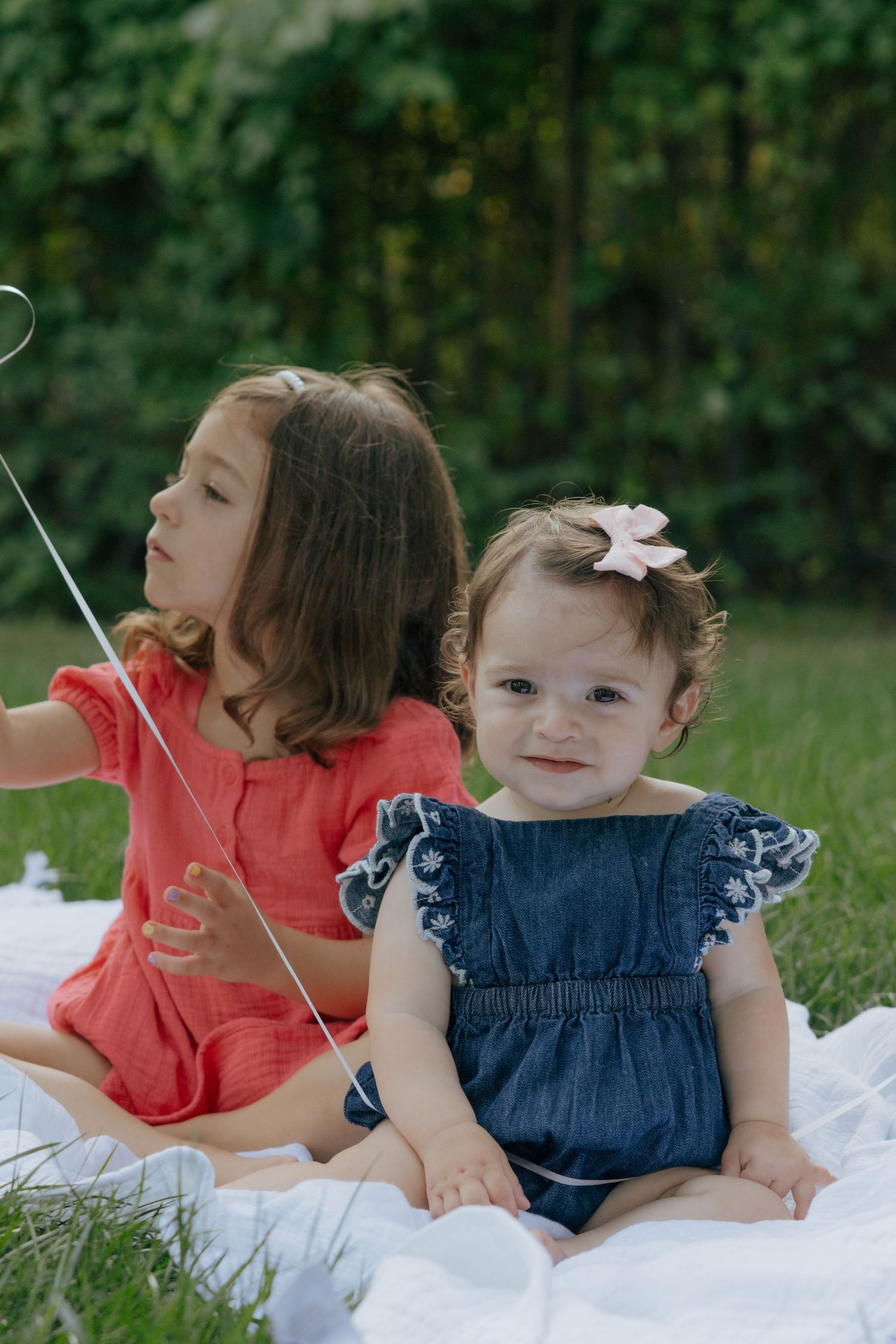 Two children in coral and denim outfits sitting together on a white blanket in a garden setting.