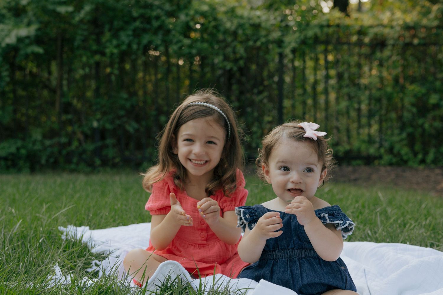 Two children in bright coral and navy blue dresses sitting on a white blanket against a green backdrop.