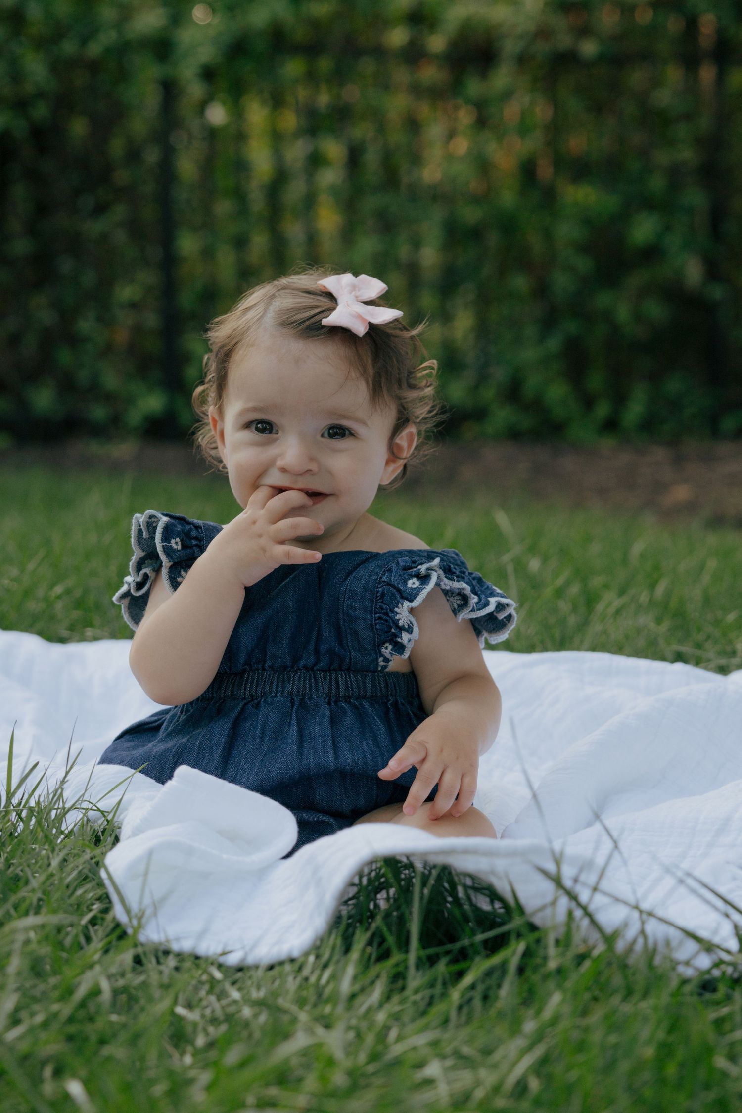Small child in denim dress with fluttered sleeves sitting on white blanket outdoors.