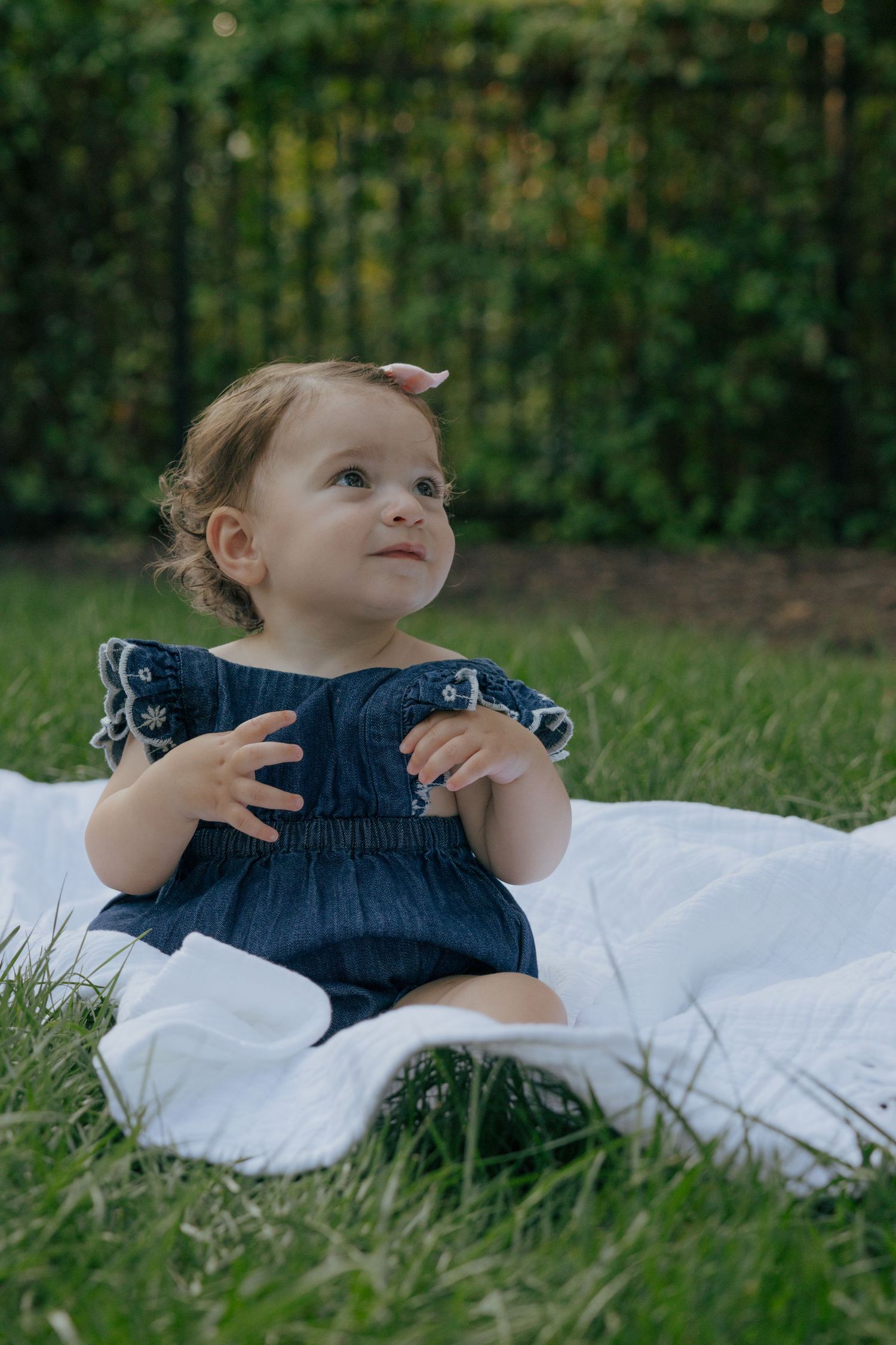 Young child in navy blue dress looking up while sitting on white blanket in garden setting.
