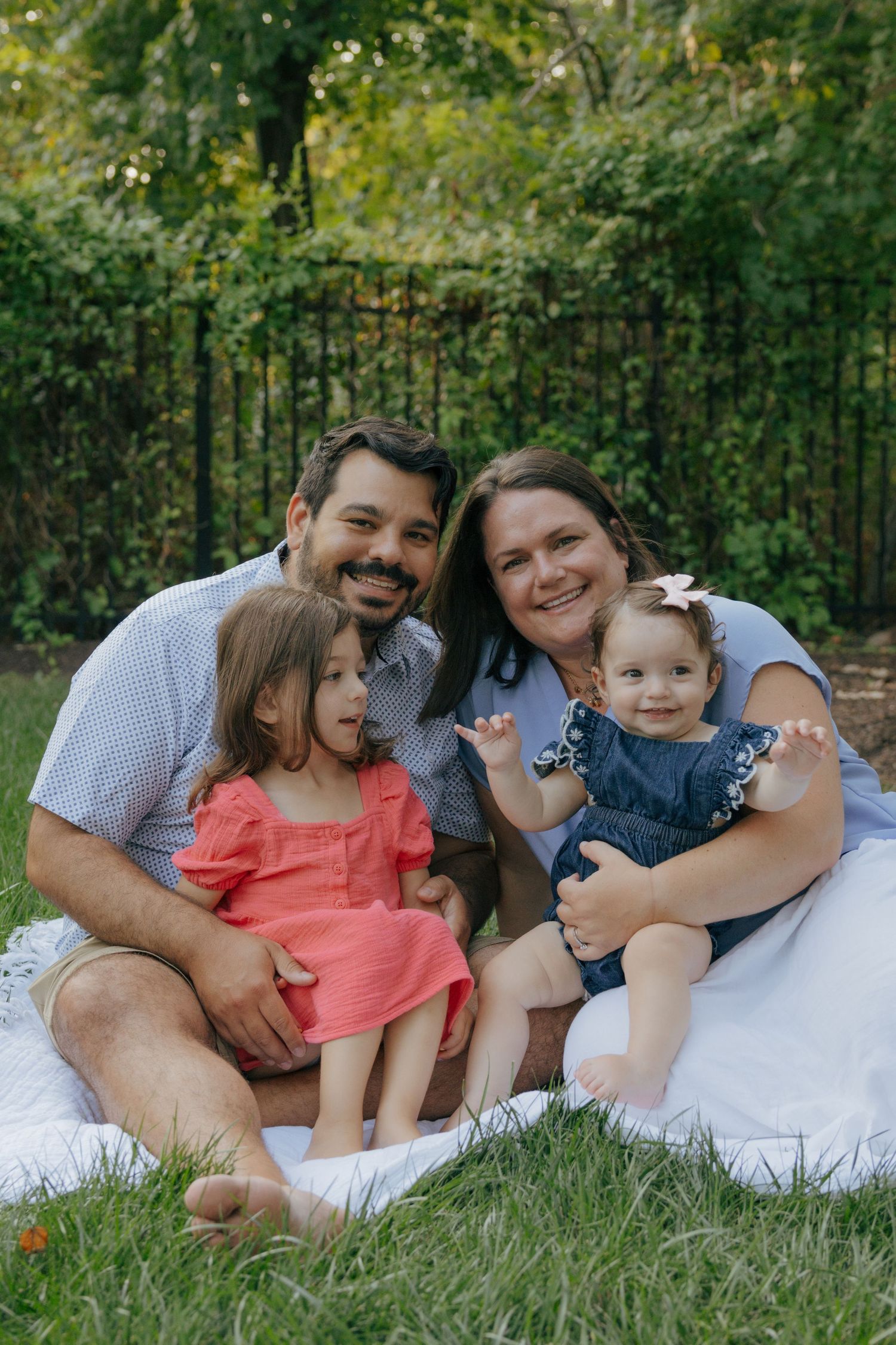 Family of four sitting together on white blanket during outdoor photo session.