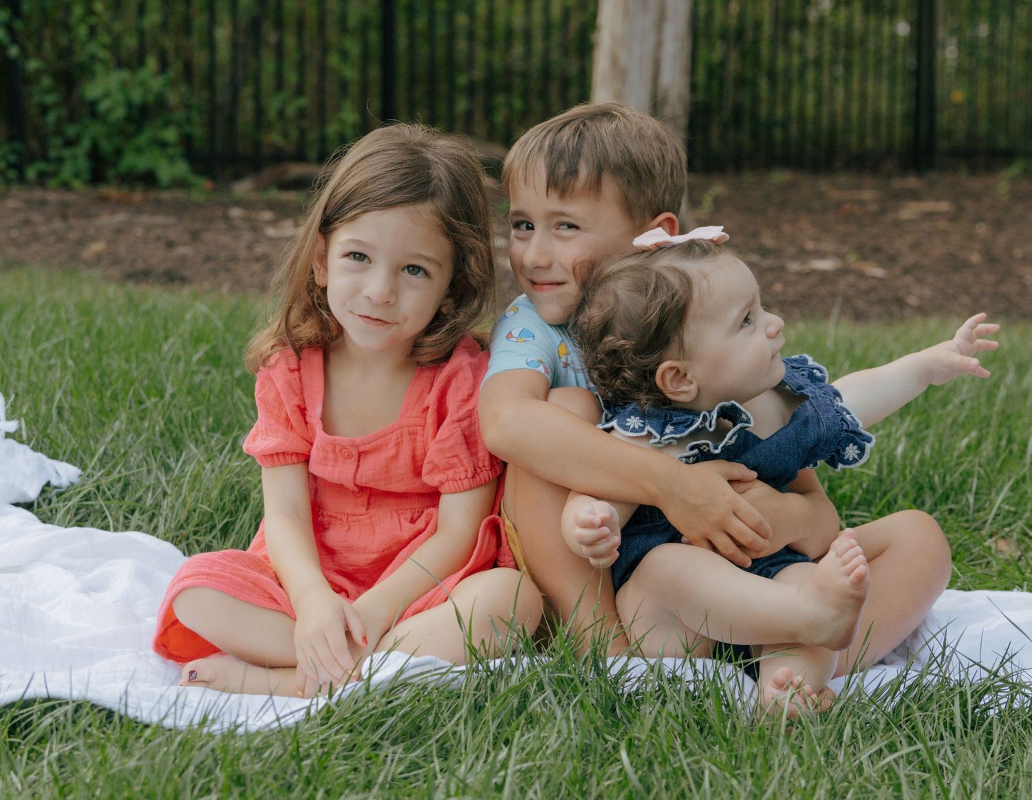 Three children sitting close together on white blanket during outdoor photo session.