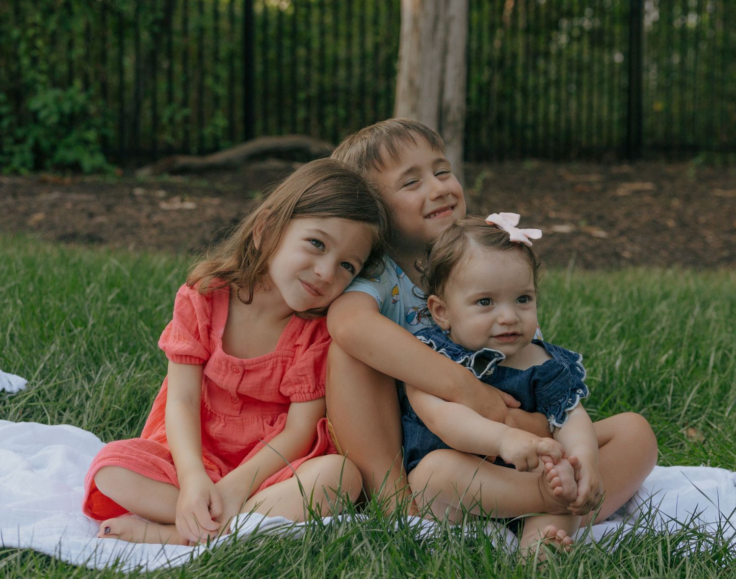 Three children cuddling together on white blanket during garden photo session.