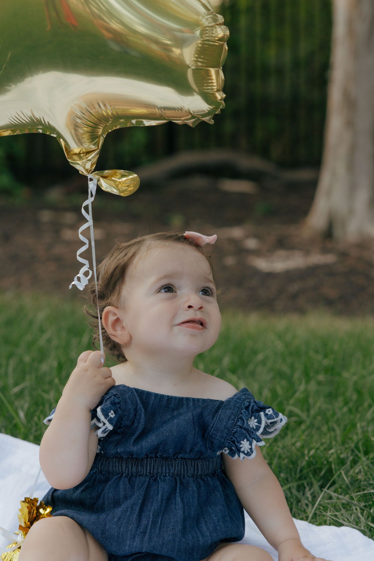 Toddler in blue dress looks up at gold balloon during outdoor cake smash photo session.