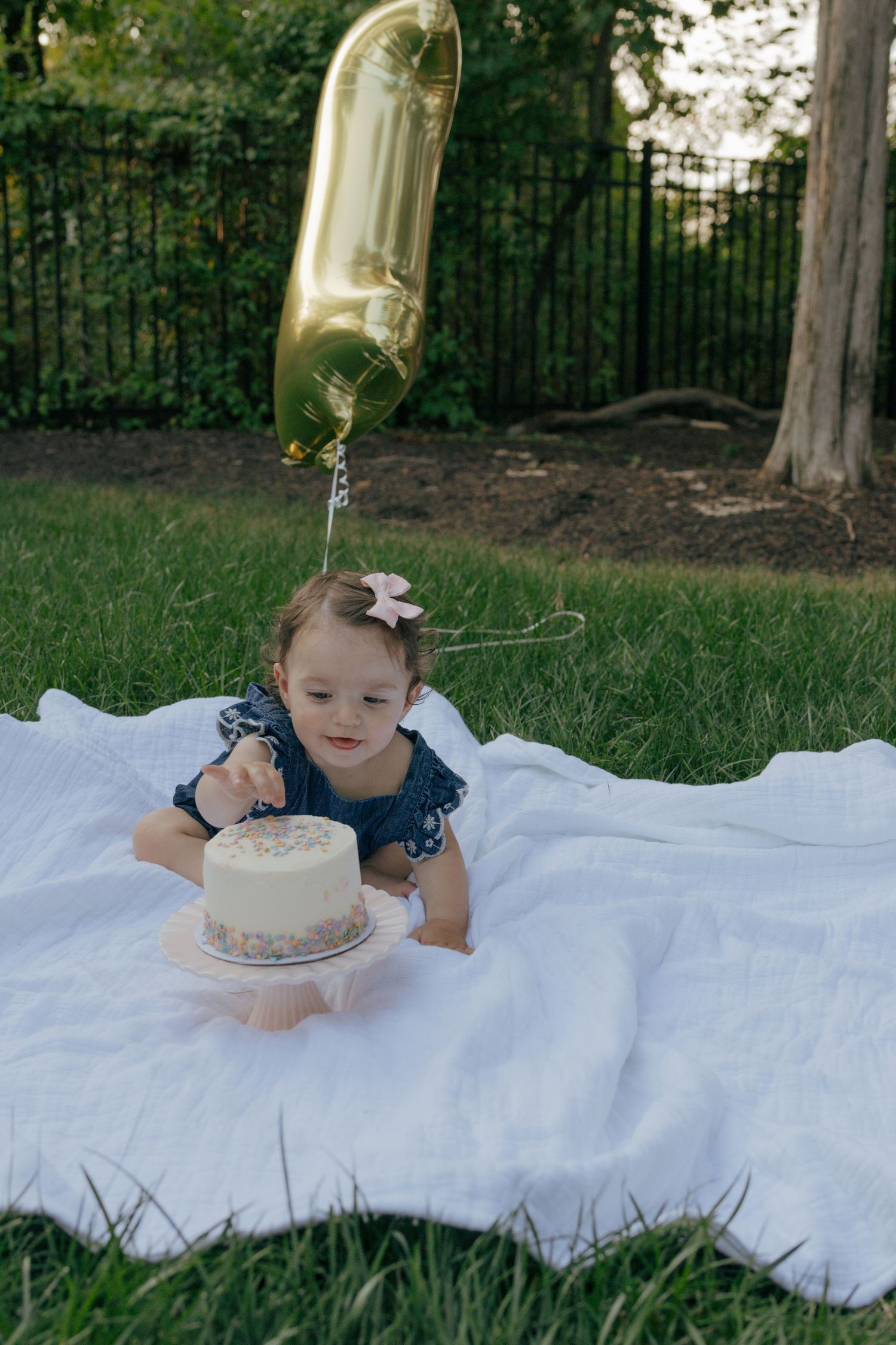 Baby in denim dress reaches for white cake during outdoor celebration.