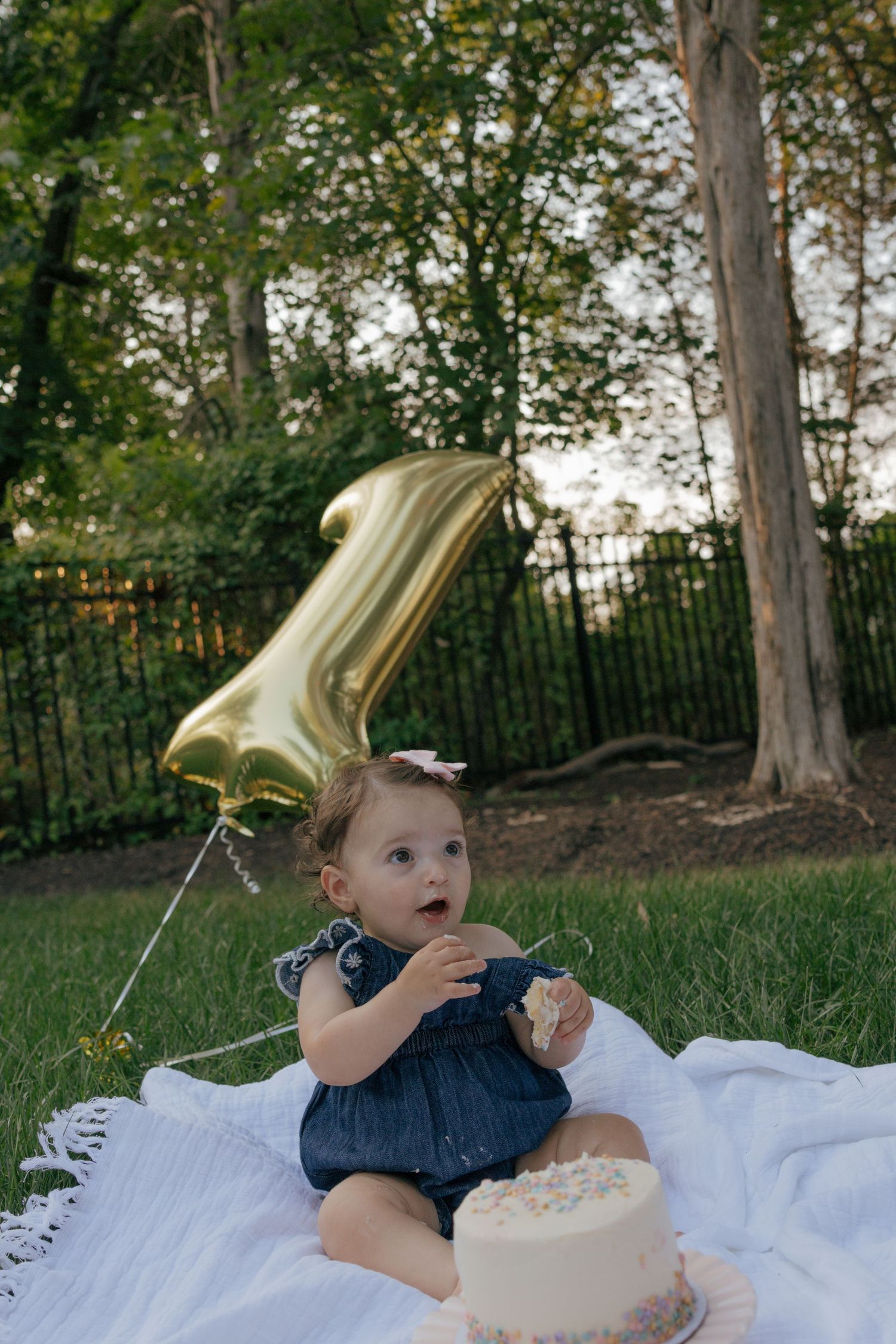 Child in denim dress with gold balloon reaches for white cake outdoors.