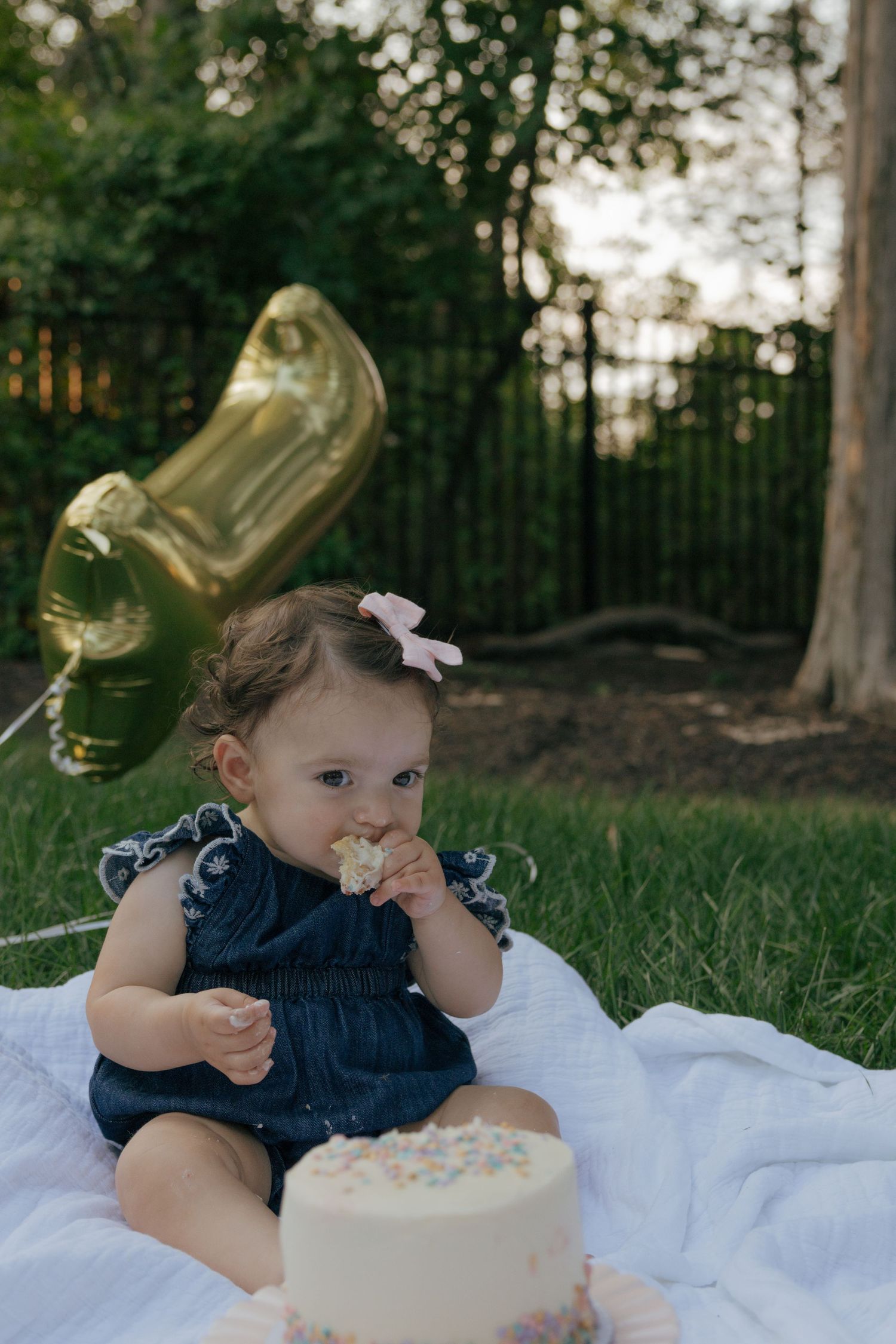 Baby in denim dress enjoys smashing cake during outdoor photo session with gold balloon and white blanket.