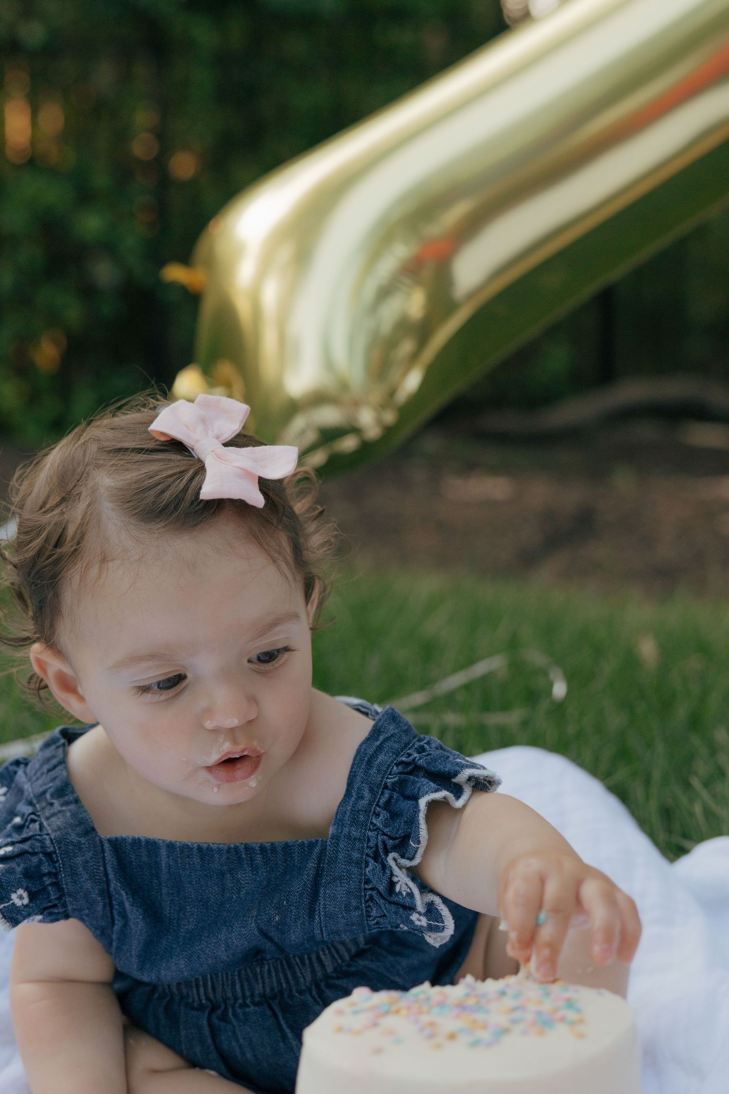 Baby wearing blue denim dress reaching for white cake during outdoor celebration.