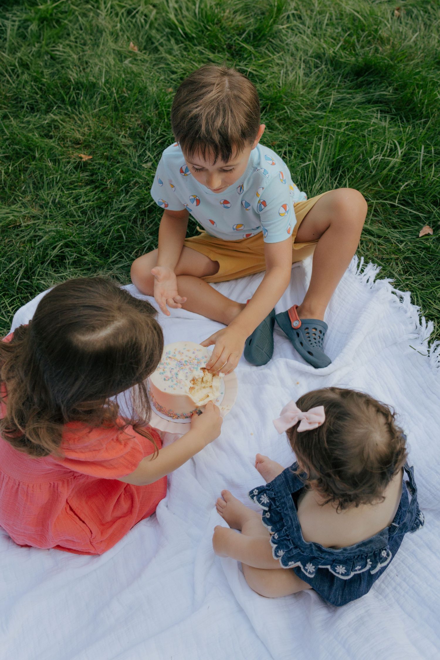 Children gather on white blanket outdoors to share cake together.