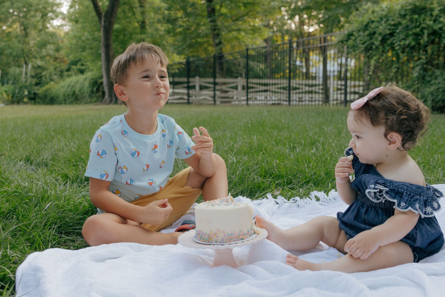 Two kids sharing snacks while sitting on a picnic blanket in a park.