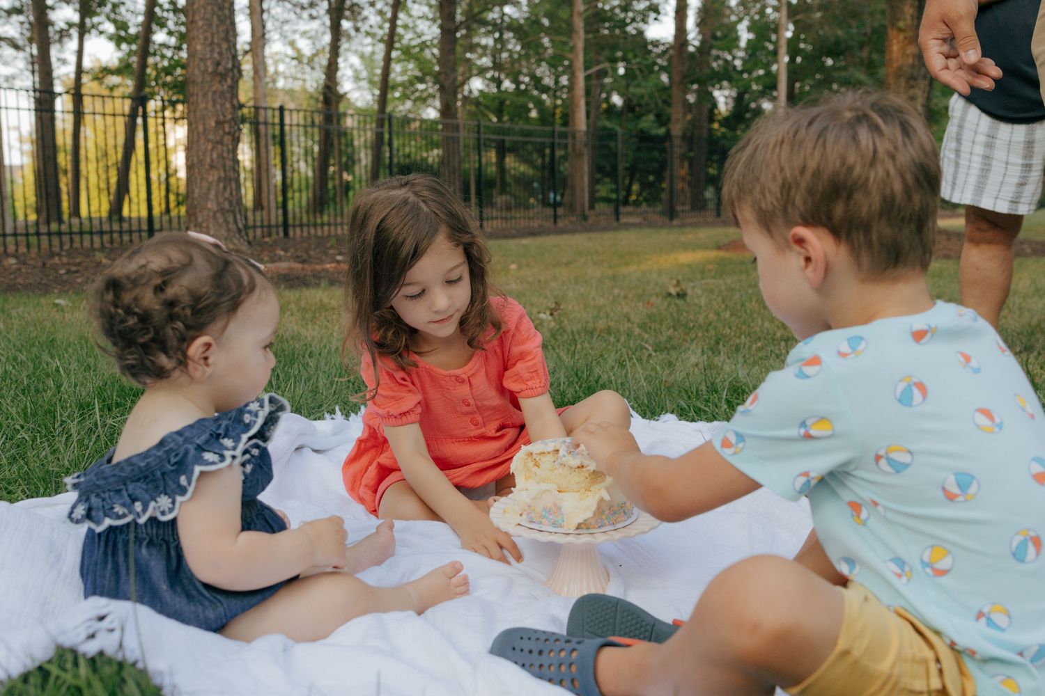 Three children having a summer picnic together on a white blanket surrounded by pine trees.