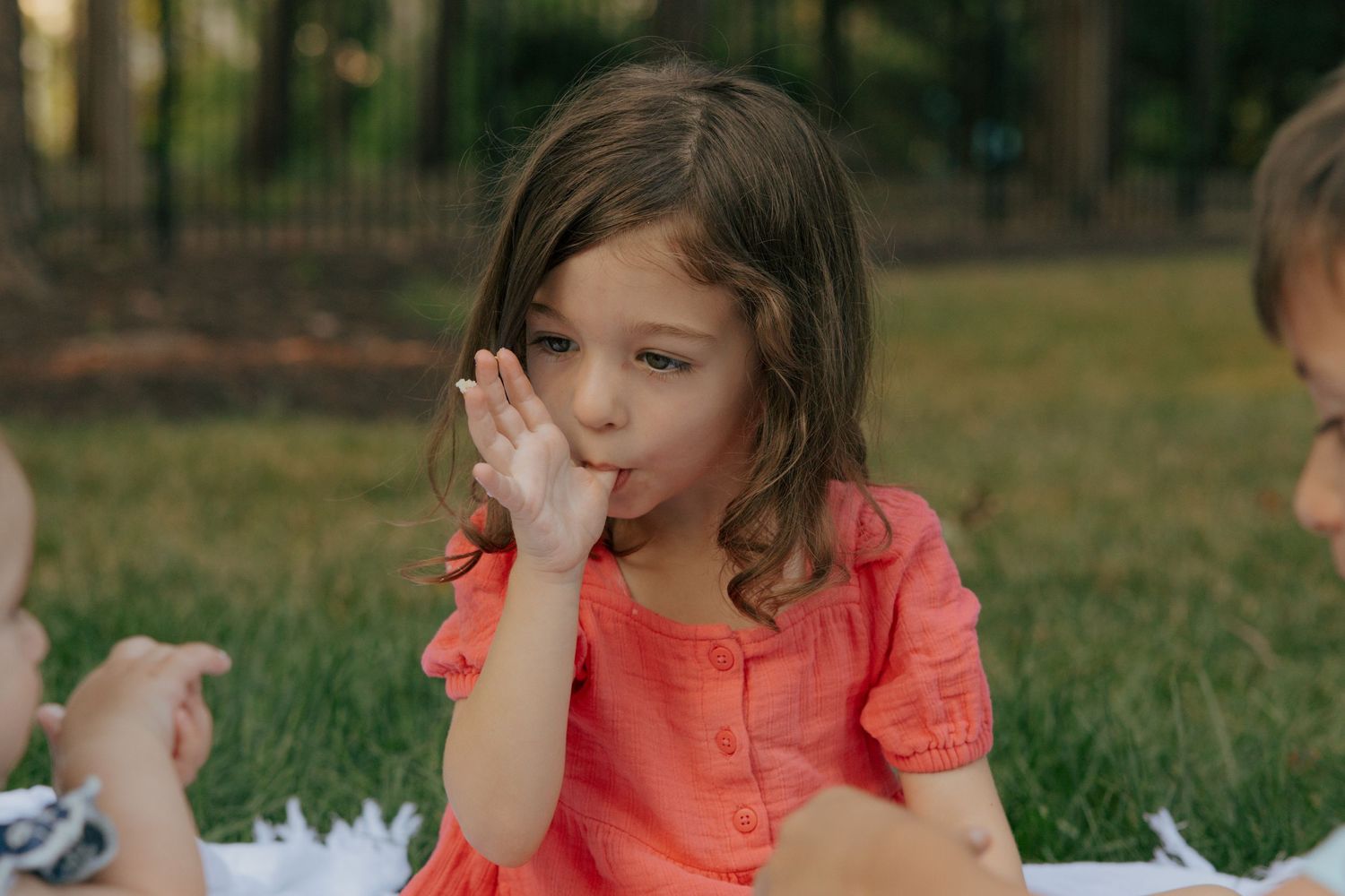 Young person in coral colored shirt eating during an outdoor picnic.