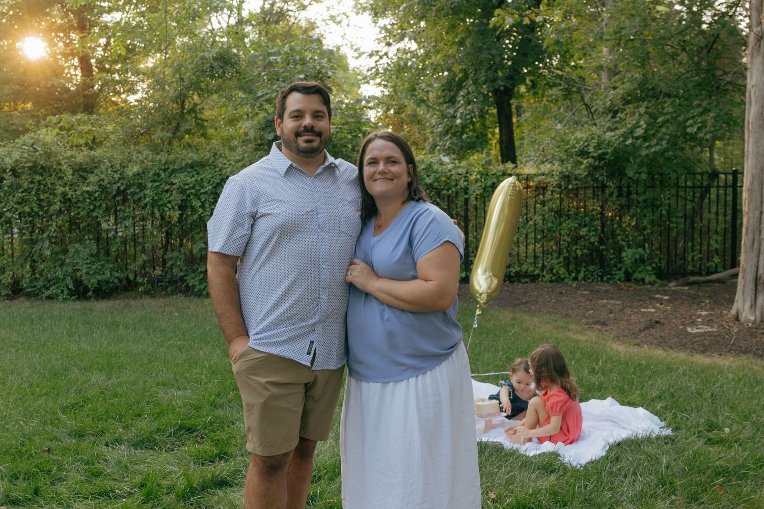Couple standing in backyard at sunset while children have picnic in background.
