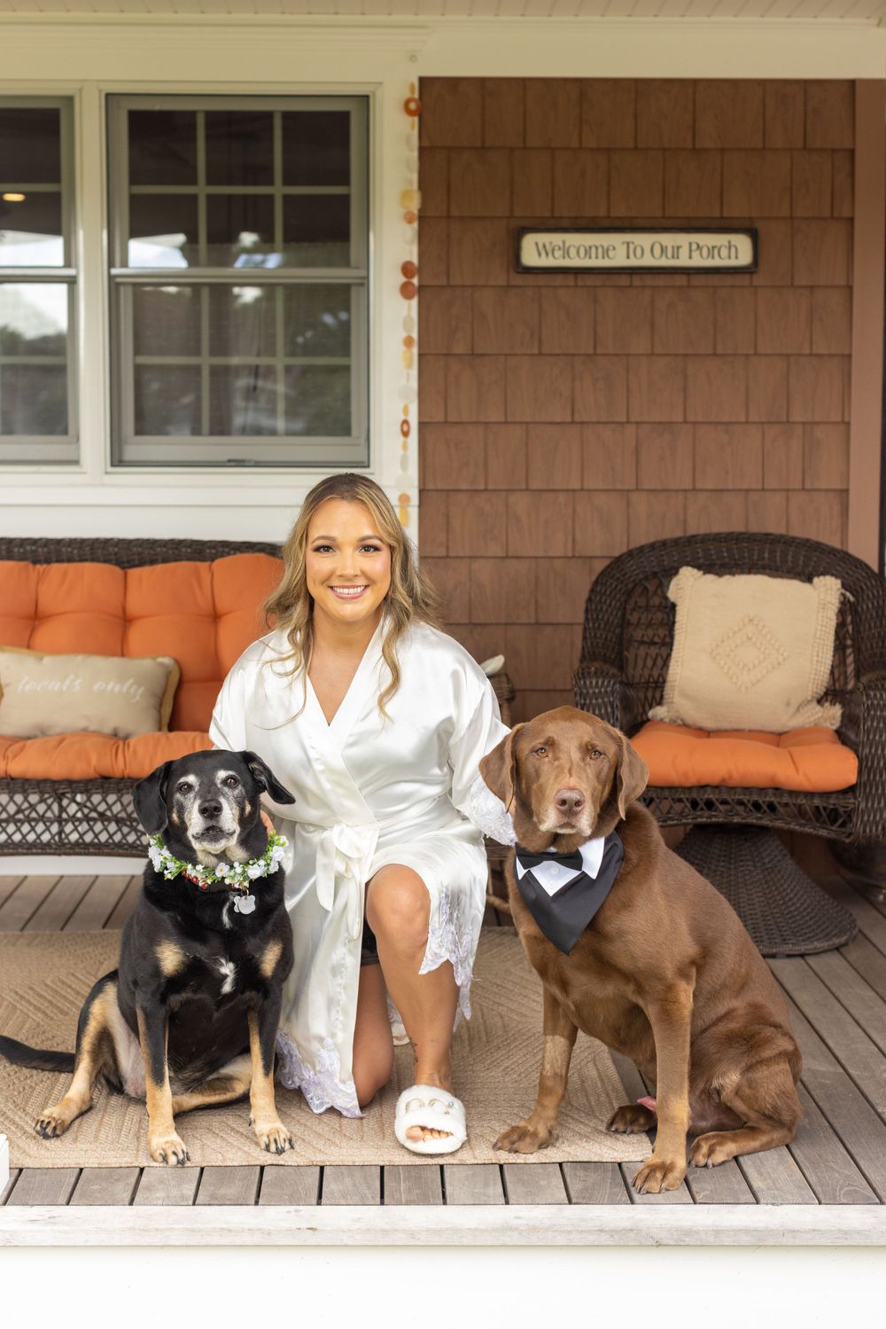 Bride in white robe sits on a porch with two dressed-up dogs, a black lab and a chocolate lab.