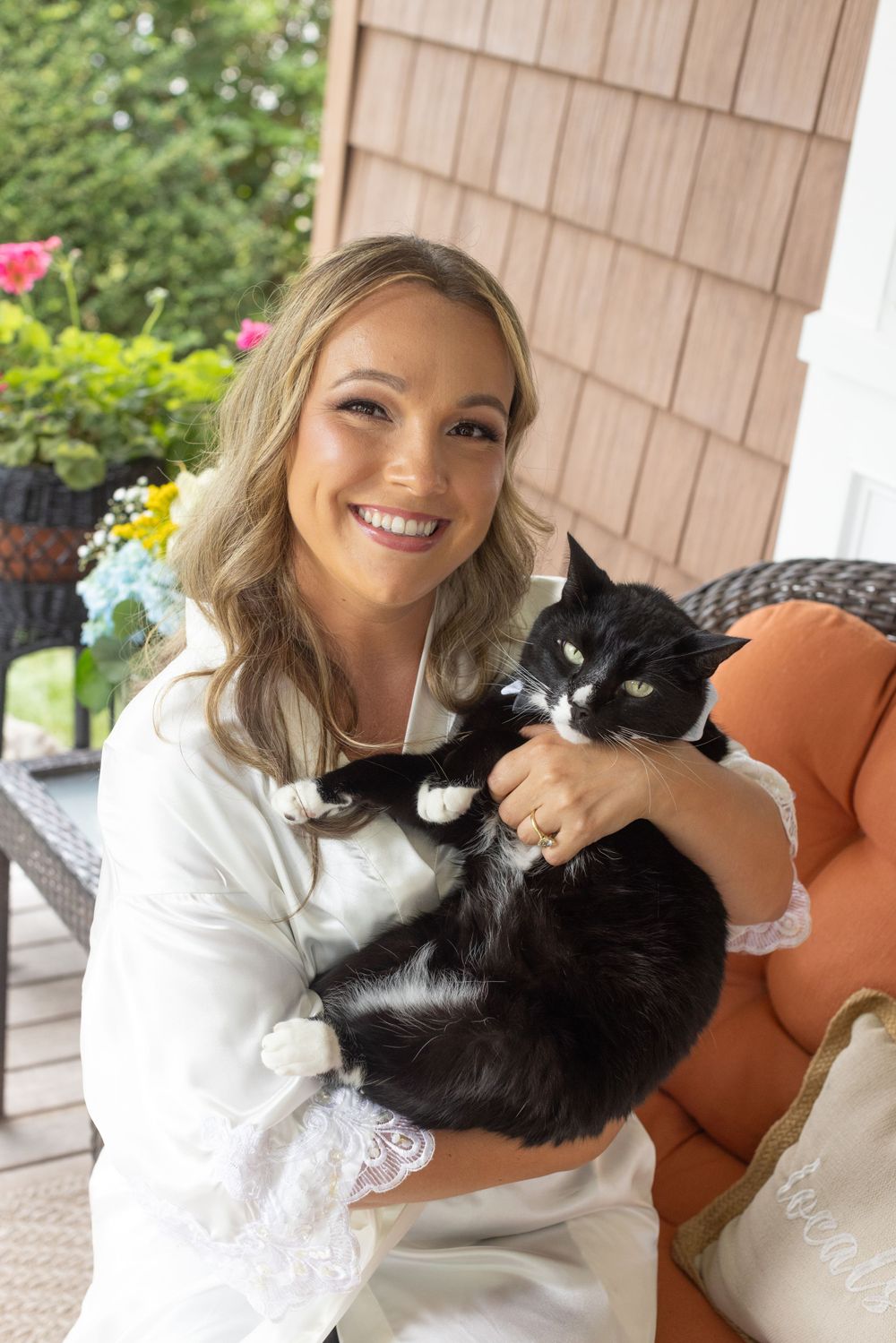 Smiling bride in white robe holds a black and white tuxedo cat on a decorated outdoor porch.