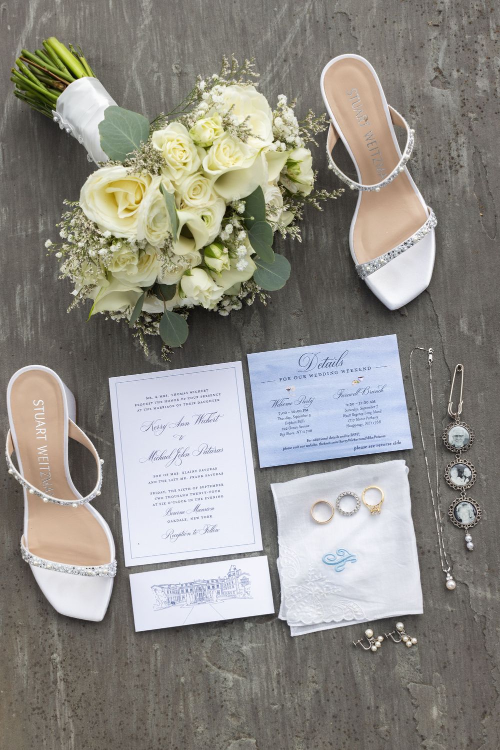 Flat lay of wedding details including invitations, rings, white heels, bouquet, and jewelry on gray surface.