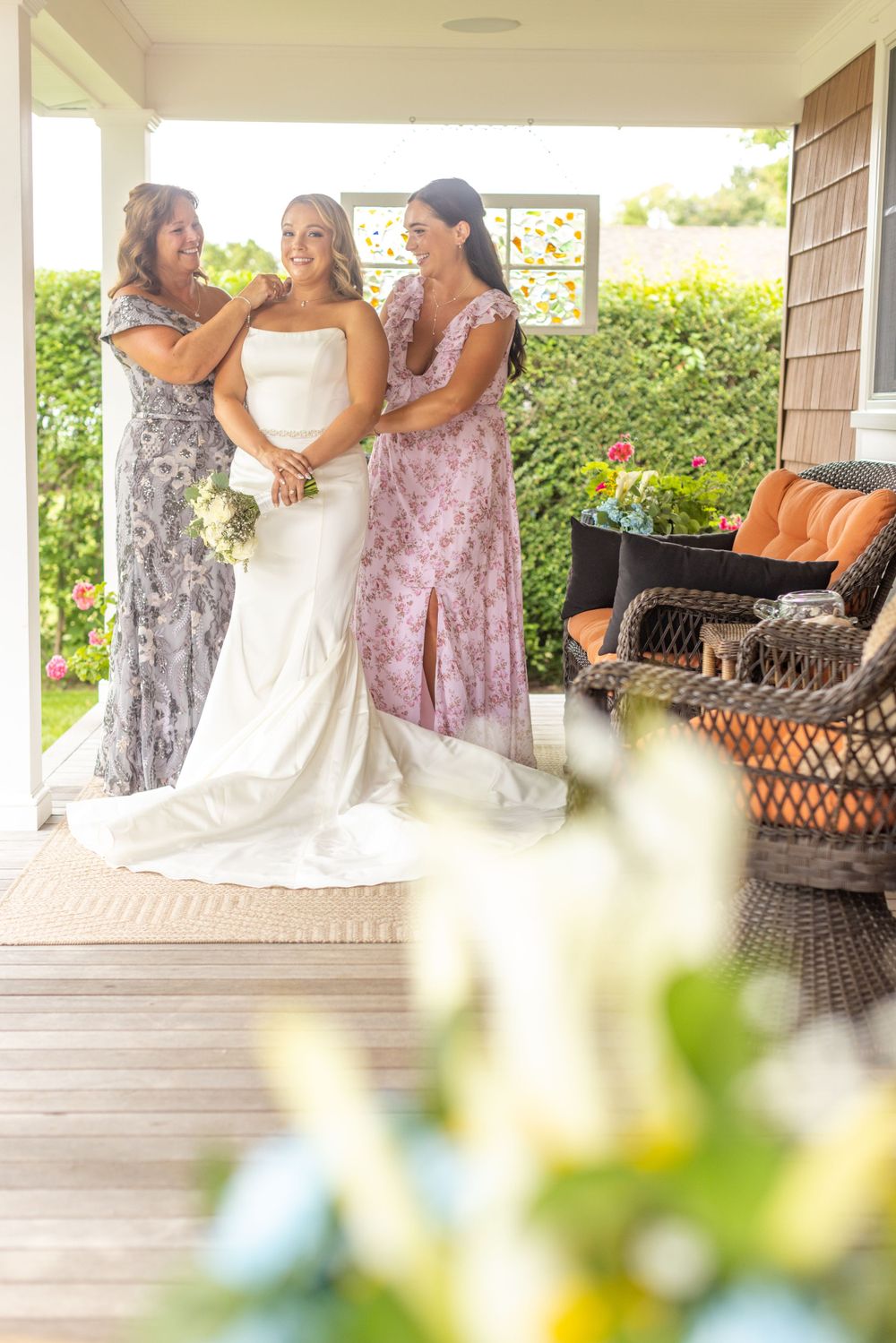 Bride in white gown holds bouquet while two women in floral dresses assist her on a sunny porch.