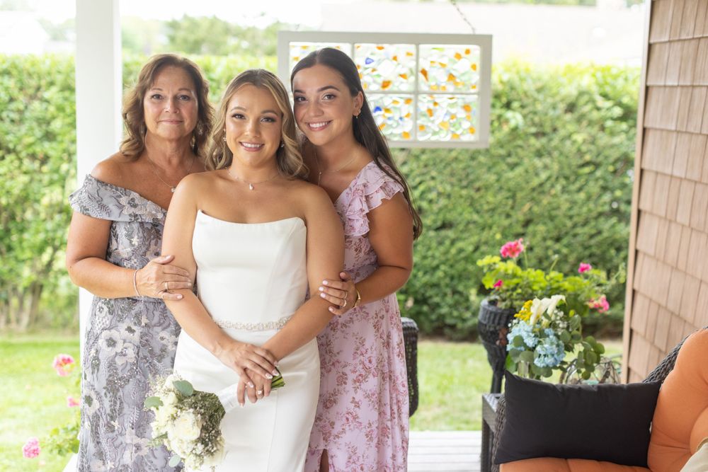 Bride in strapless white gown poses with two smiling women in floral dresses on a garden porch.