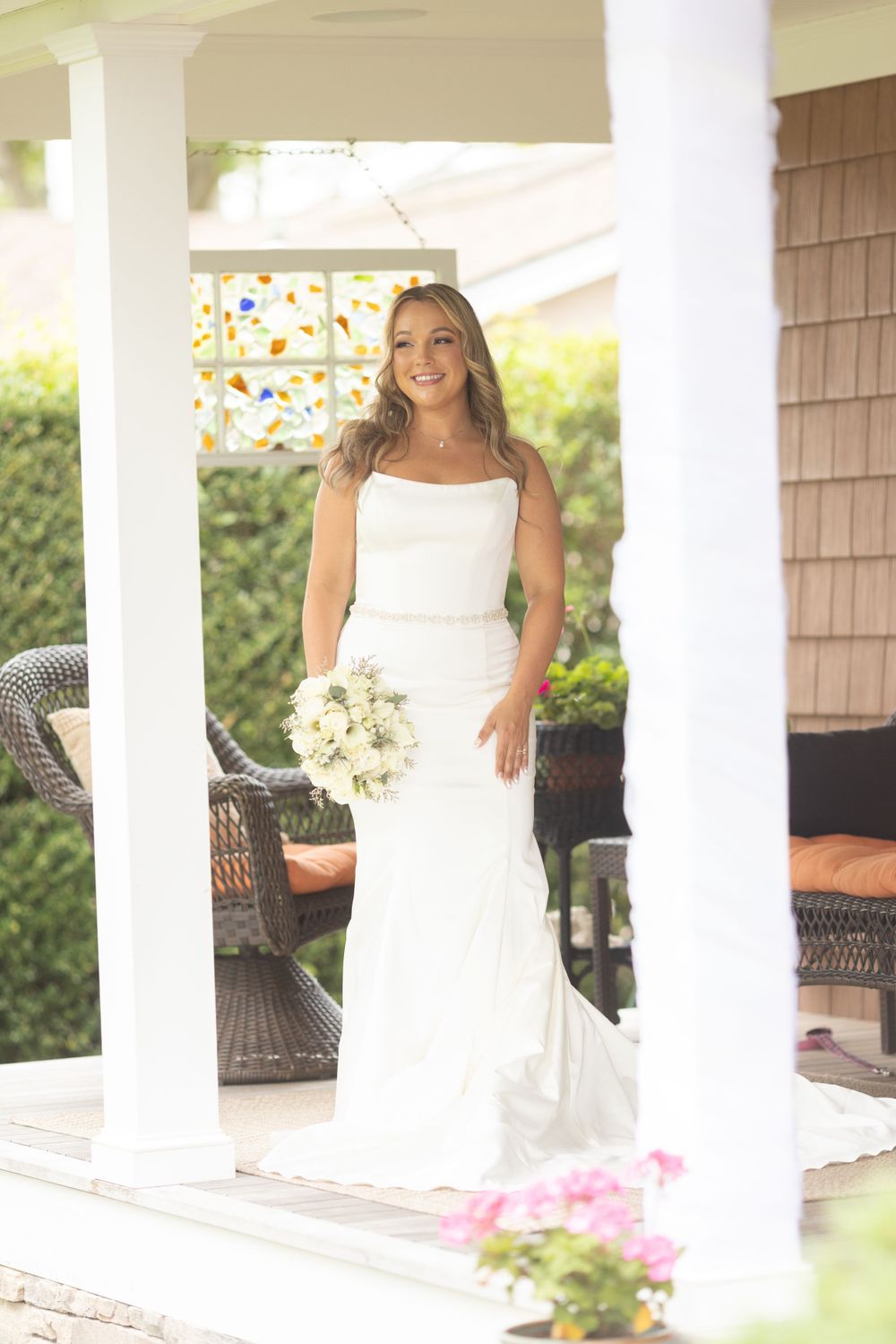 Elegant bride in white mermaid gown holds a white bouquet while standing between white porch columns.