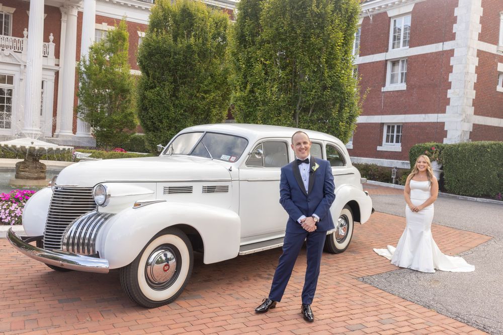 Groom stands proudly by a vintage white classic car as bride poses gracefully in the background.