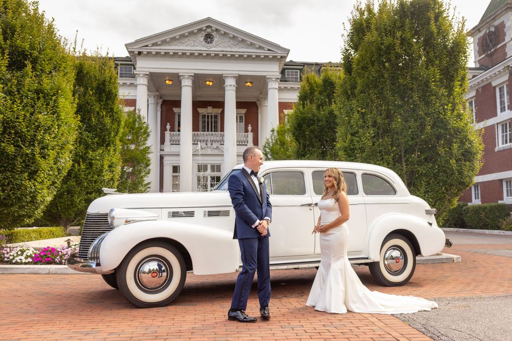 Bride and groom stand together in front of a grand white mansion beside a classic vintage white car.