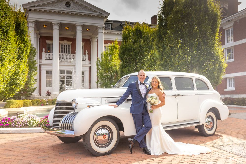 Bride and groom posing beside a classic white vintage car in front of an elegant mansion venue.