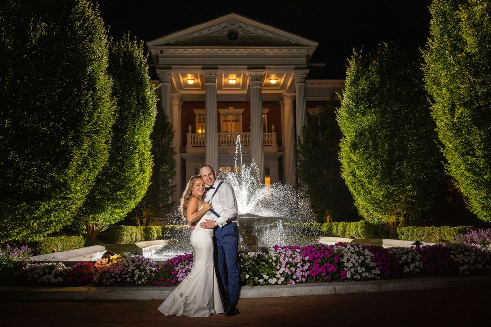Newlyweds pose romantically in front of glowing fountain flanked by tall hedges at elegant nighttime wedding venue.
