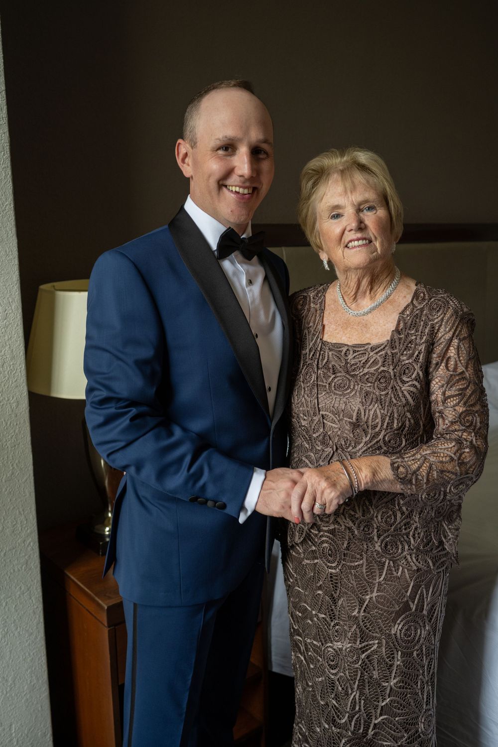 Groom in blue tuxedo poses with elderly mother holding hands before wedding ceremony.