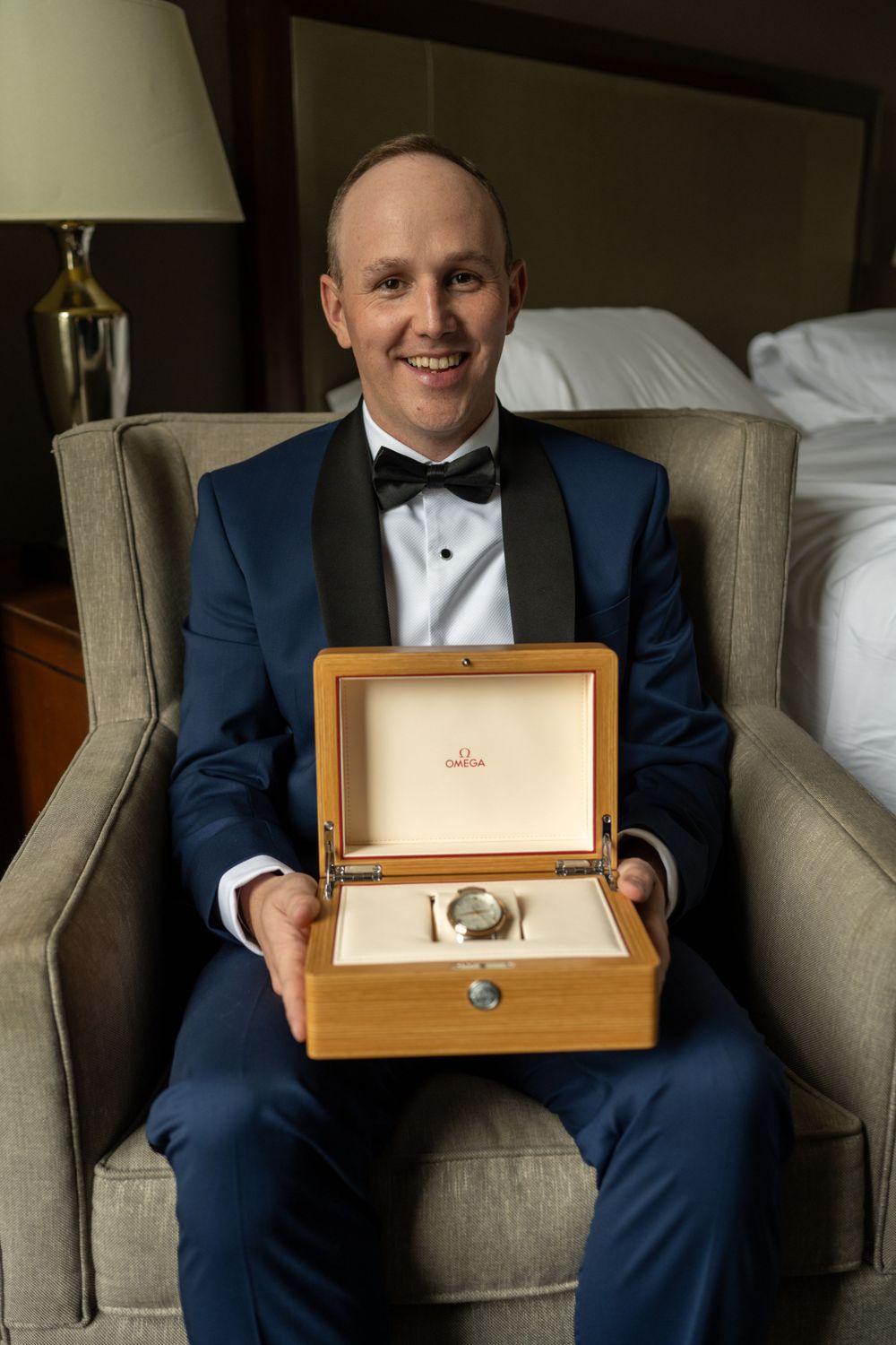 Smiling groom in blue tuxedo proudly displays Omega watch gift in wooden box.