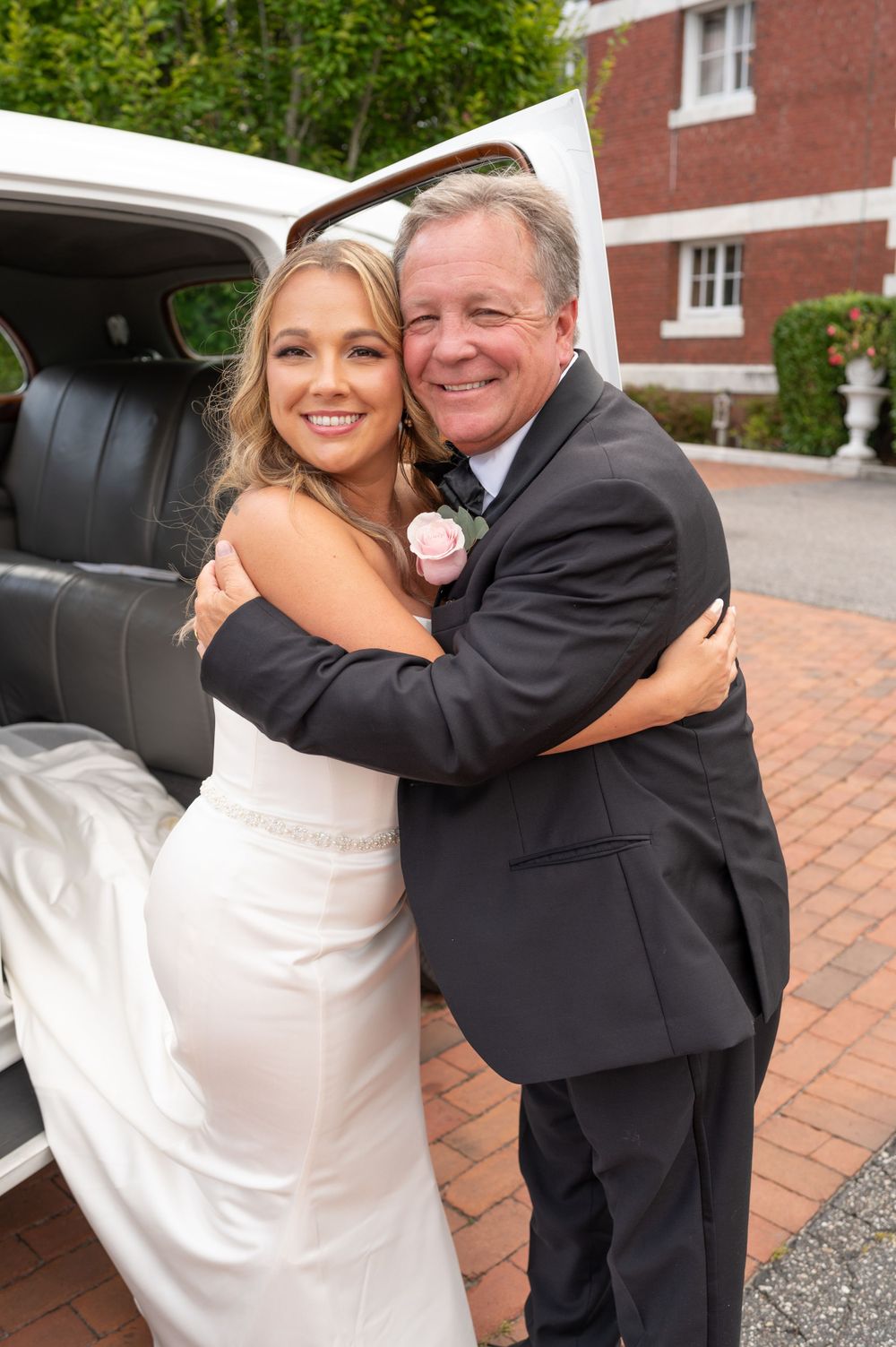Radiant bride in white gown hugs father beside white limousine before wedding ceremony.