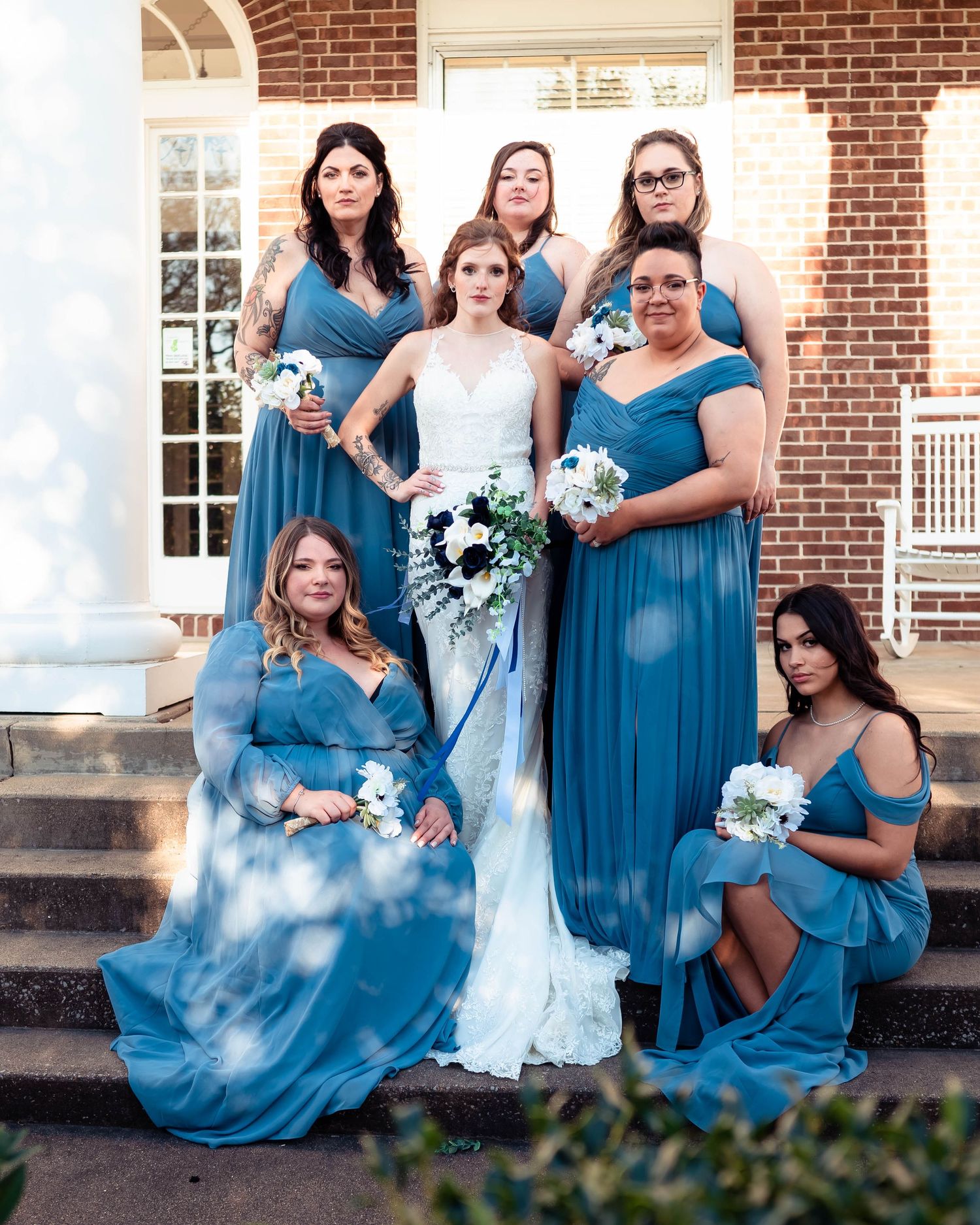 Bridesmaids in flowing blue dresses pose together against a brick wall backdrop.