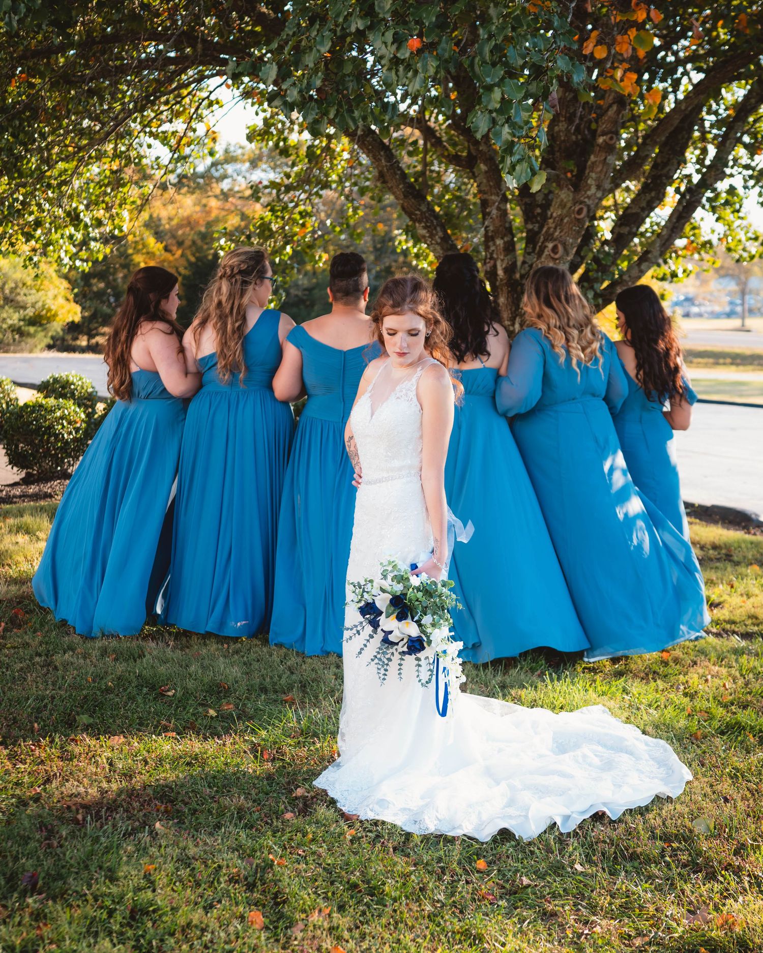 Wedding group poses on grass under trees wearing matching blue gowns.