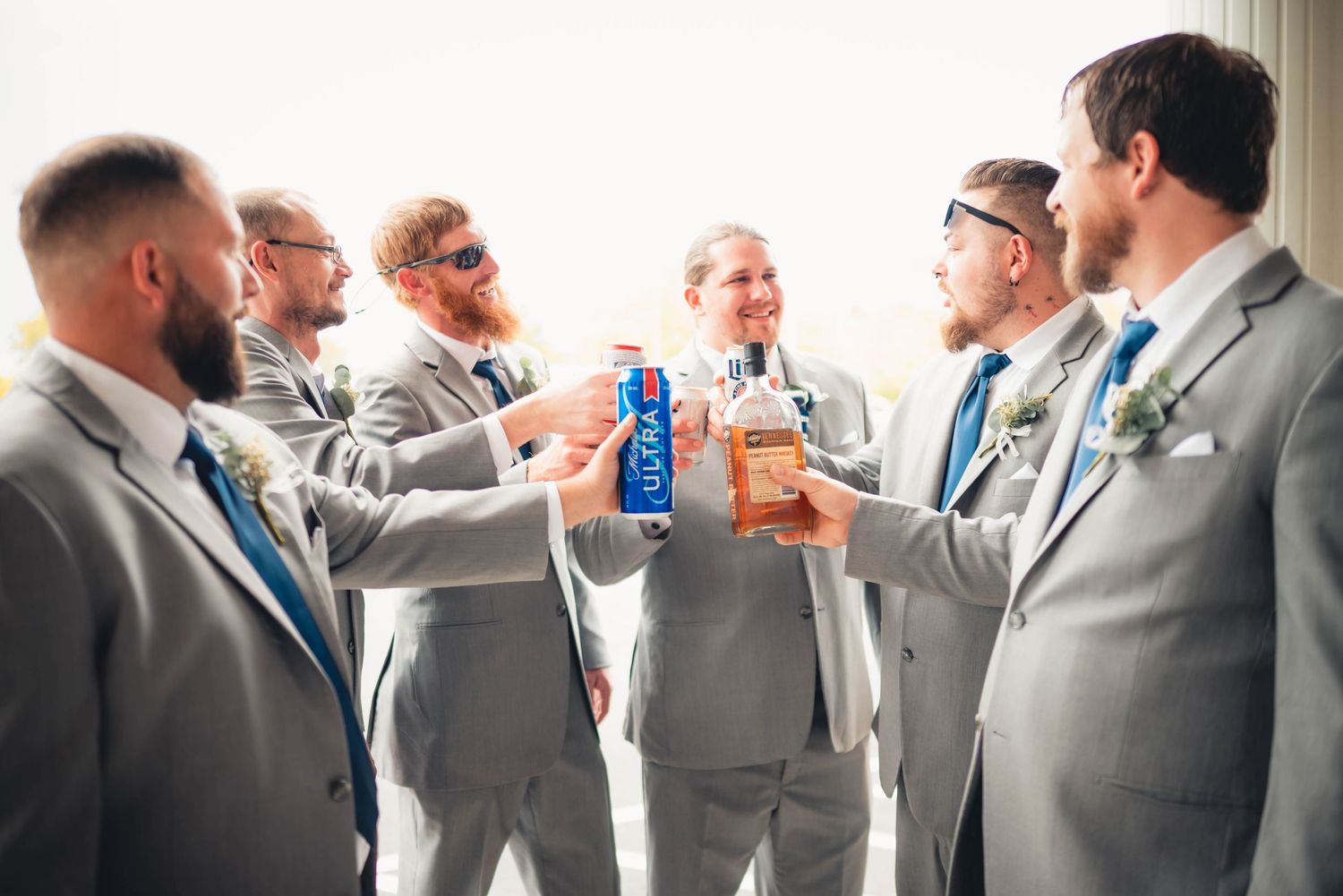 Groomsmen in matching grey suits toast with drinks outdoors.