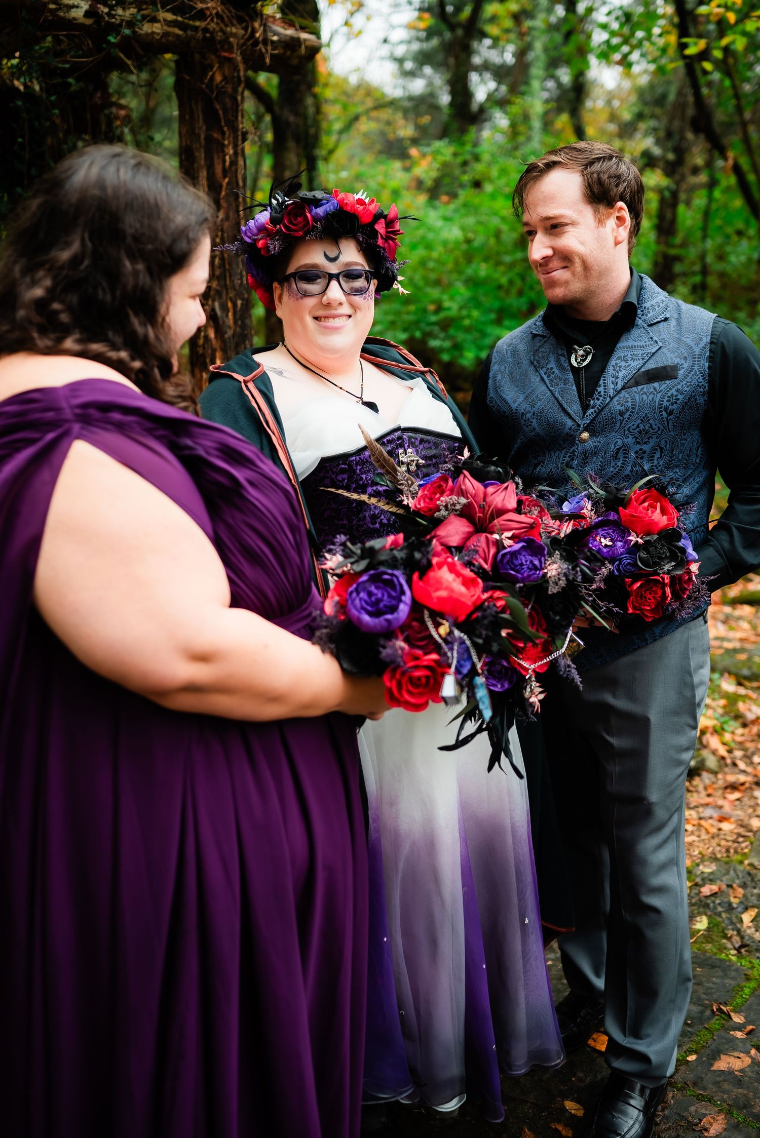 Wedding party members with deep purple dresses and vibrant red bouquets in woodland setting.
