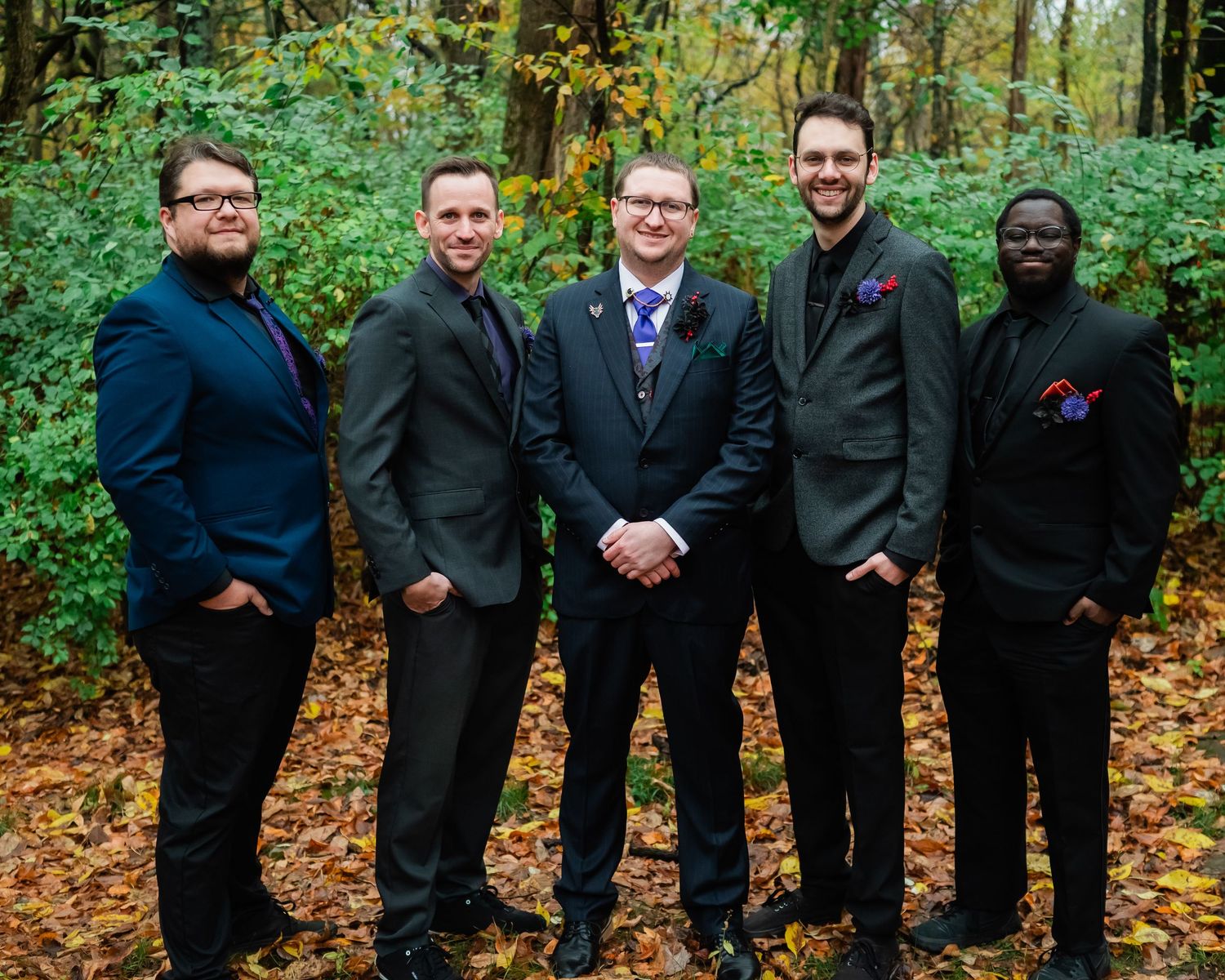 Groomsmen in black suits pose casually in wooded outdoor setting during fall.