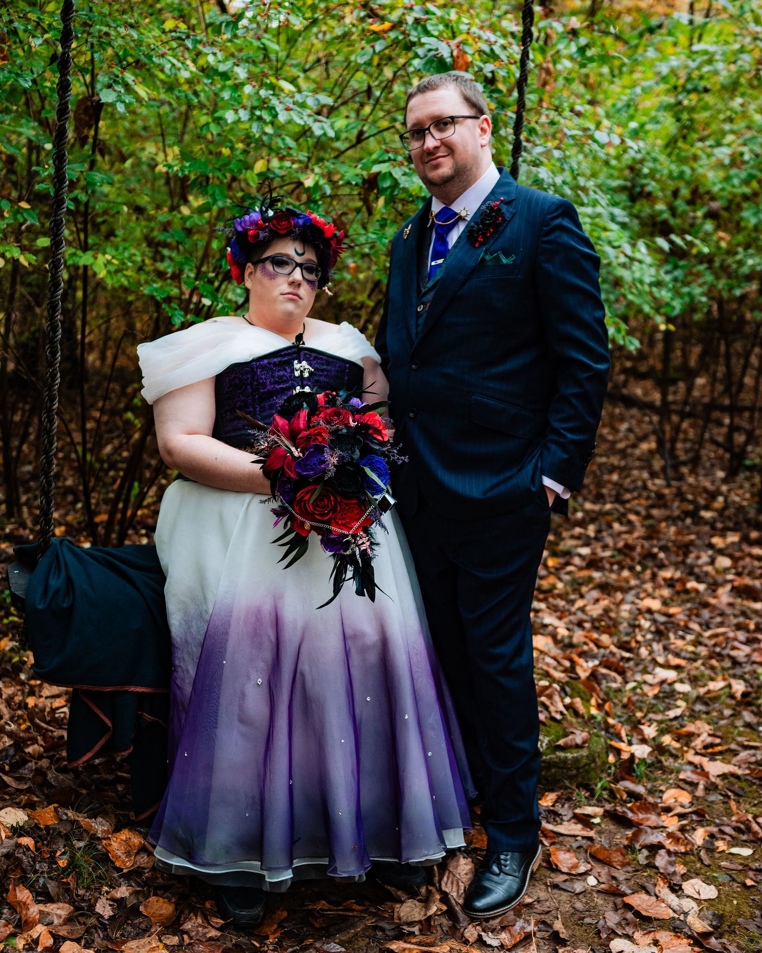 A couple in wedding attire poses in an autumn forest setting with fallen leaves and vibrant foliage.