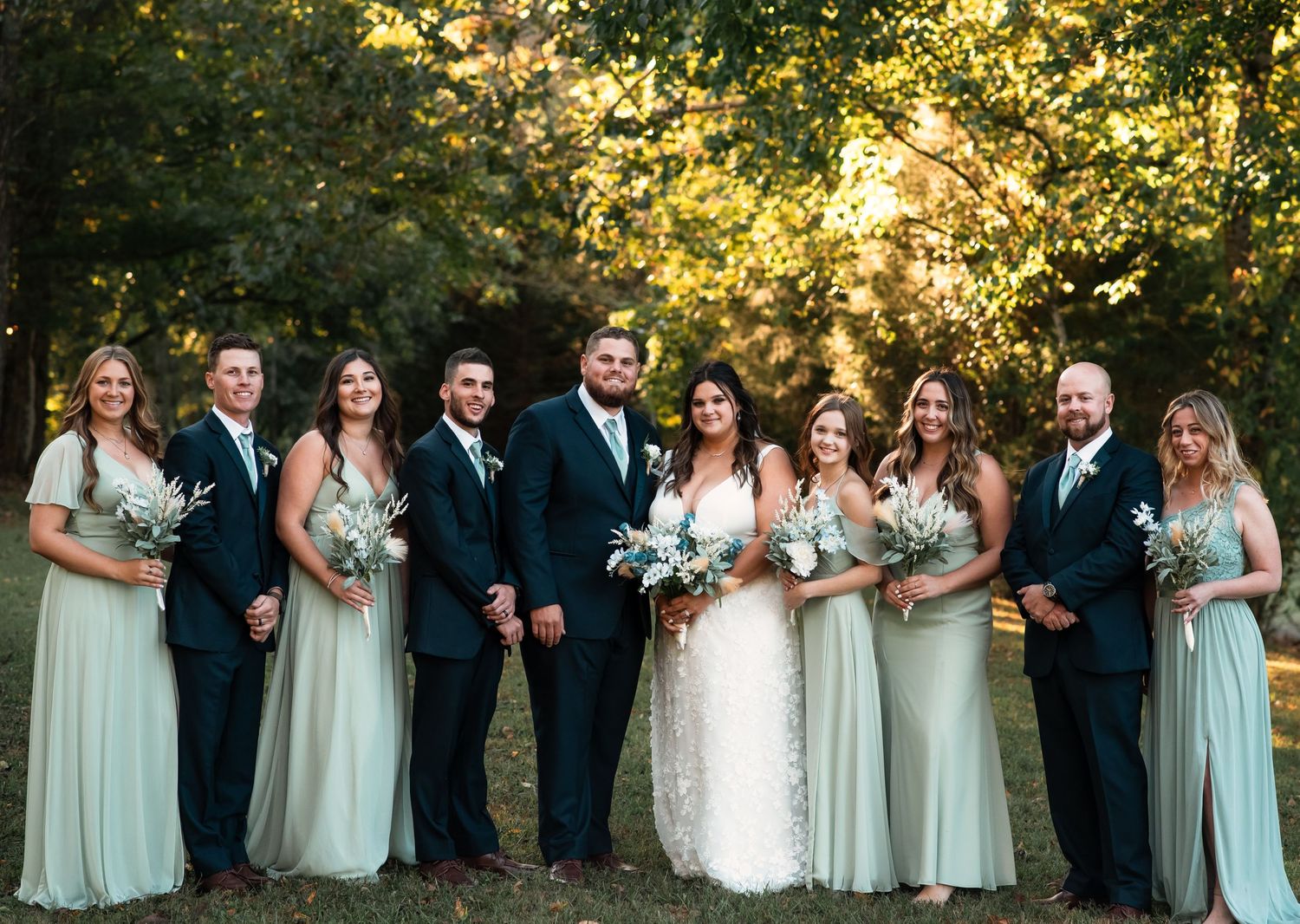 Large wedding party stands in a row against a natural backdrop with autumn foliage.