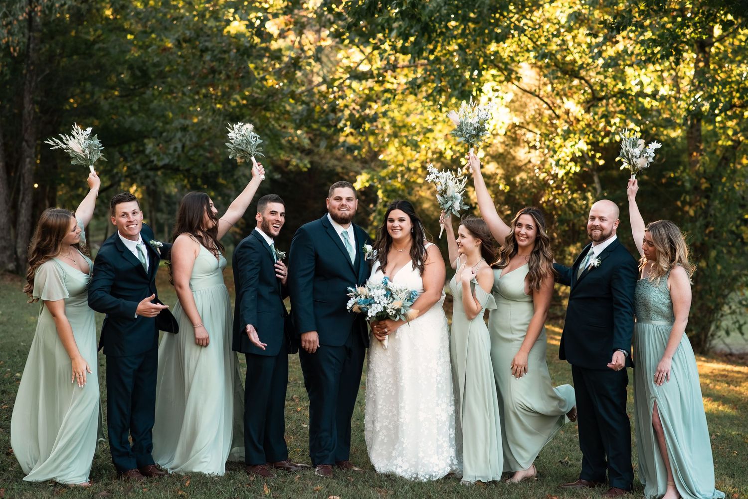 Wedding party in mint green dresses and dark suits pose together under golden sunlit trees.
