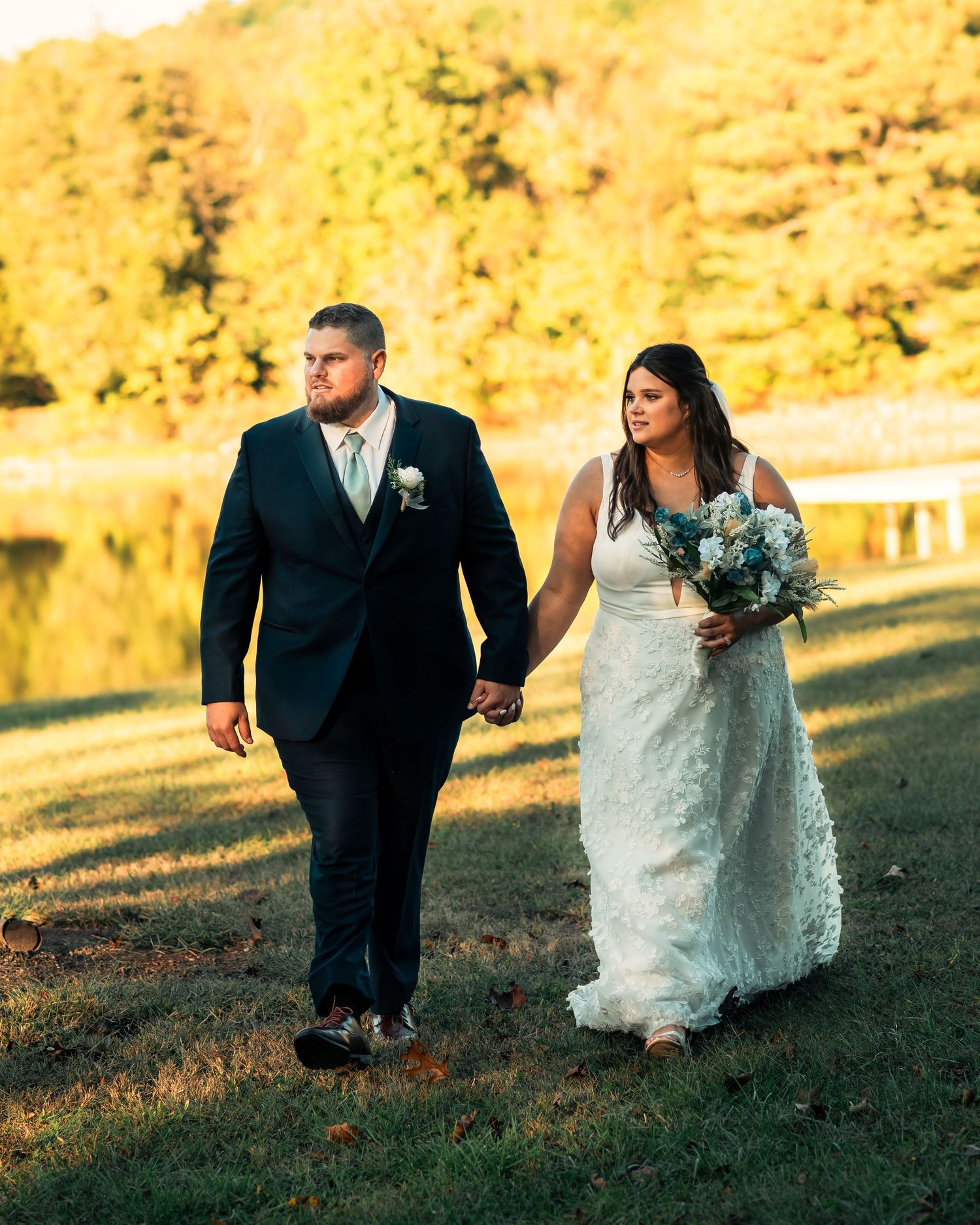 Wedding couple walking along grassy lakeside path during autumn sunset.
