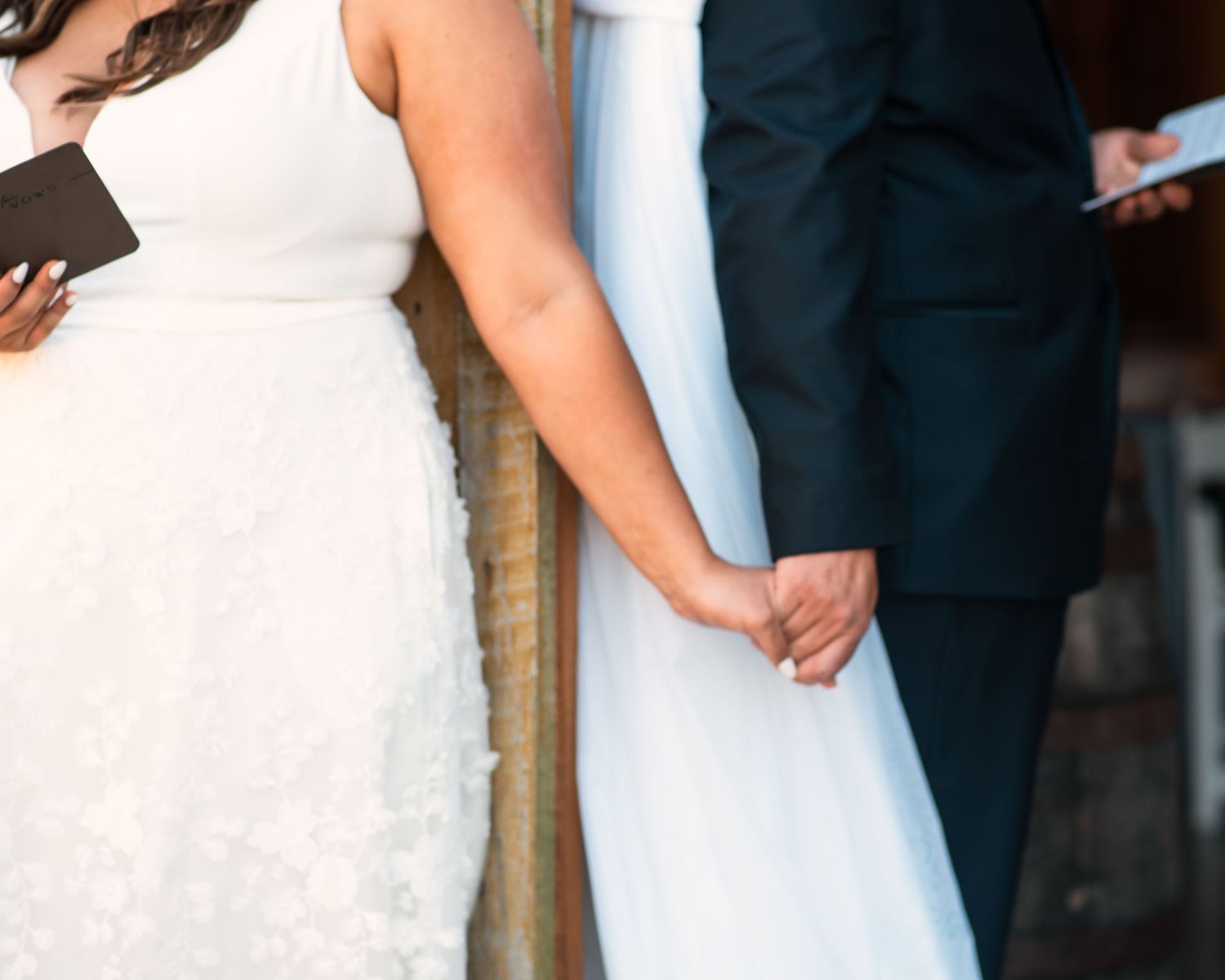 A close-up shot of a couple holding hands during their wedding ceremony, showing white dress and dark suit details.
