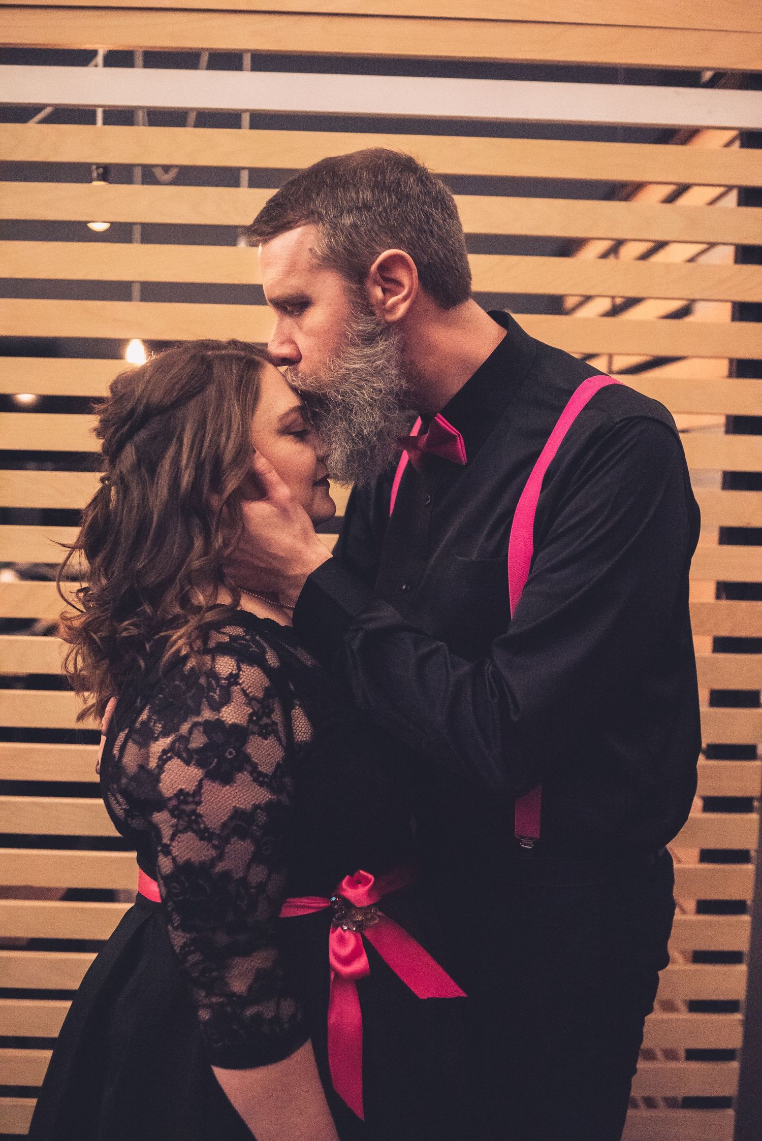 Romantic couple in black formal wear with hot pink accents embracing against wooden slat wall.