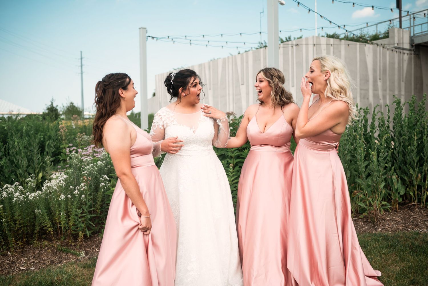 Bridesmaids in pink dresses gather outdoors under string lights for casual photos.