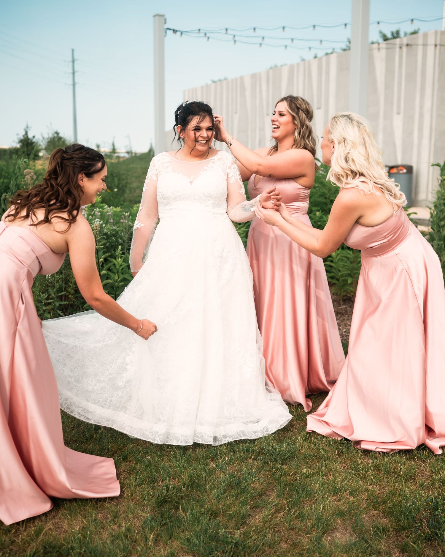 Wedding party in pink dresses poses playfully on grassy lawn with string lights overhead.