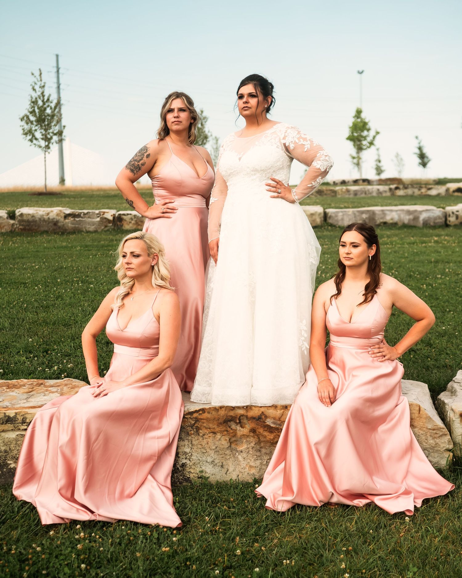 Bridesmaids in flowing pink gowns pose dramatically on outdoor stone wall.