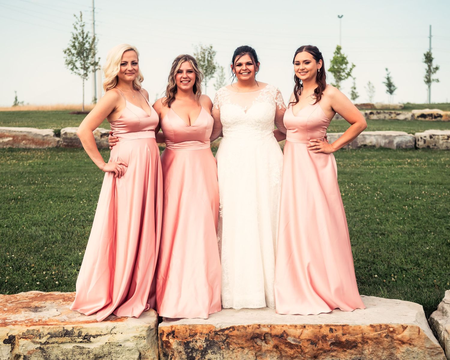 Wedding party in blush pink dresses poses together on rustic stone wall outdoors.