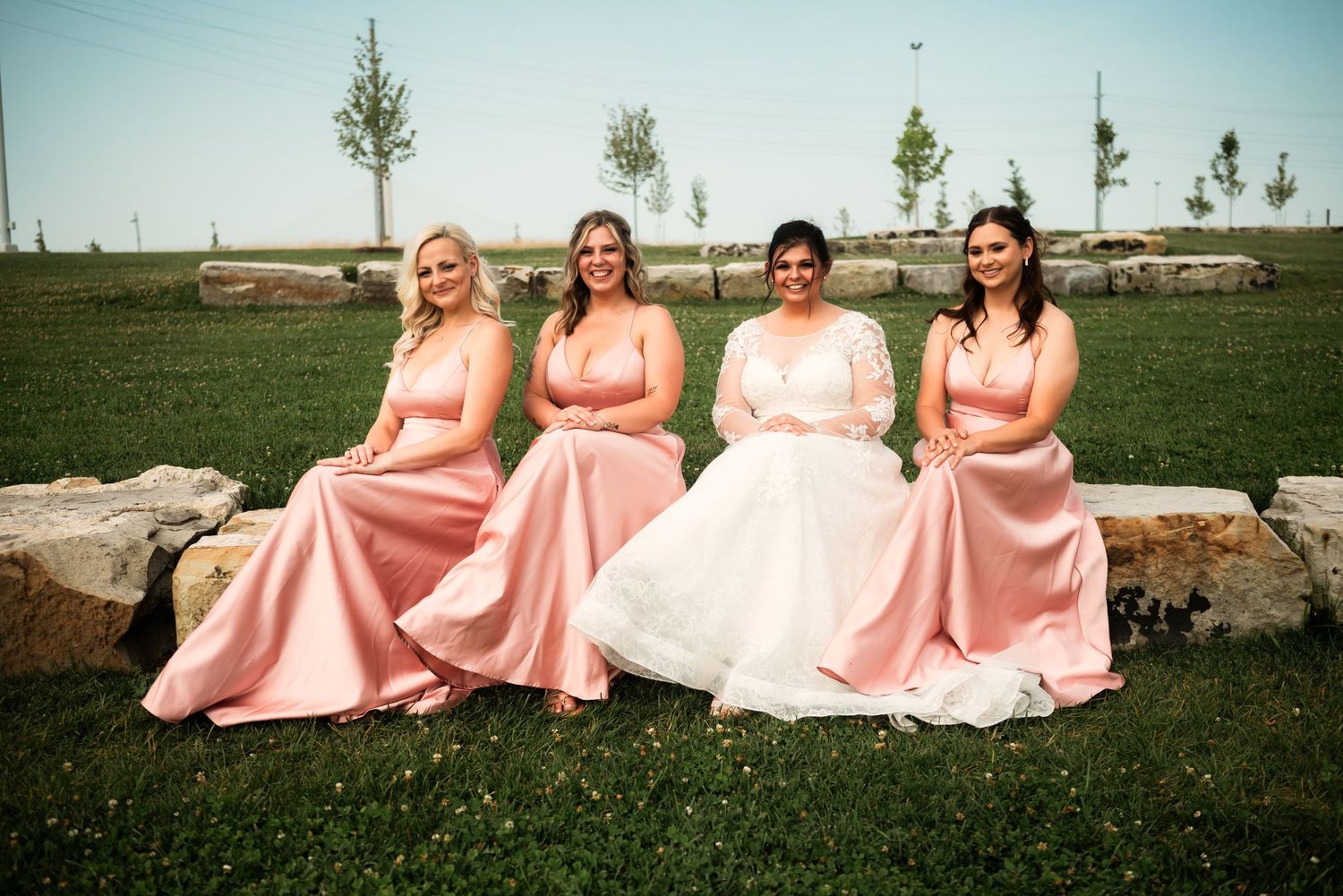 Wedding party in pink dresses sits arranged on stone steps outdoors.