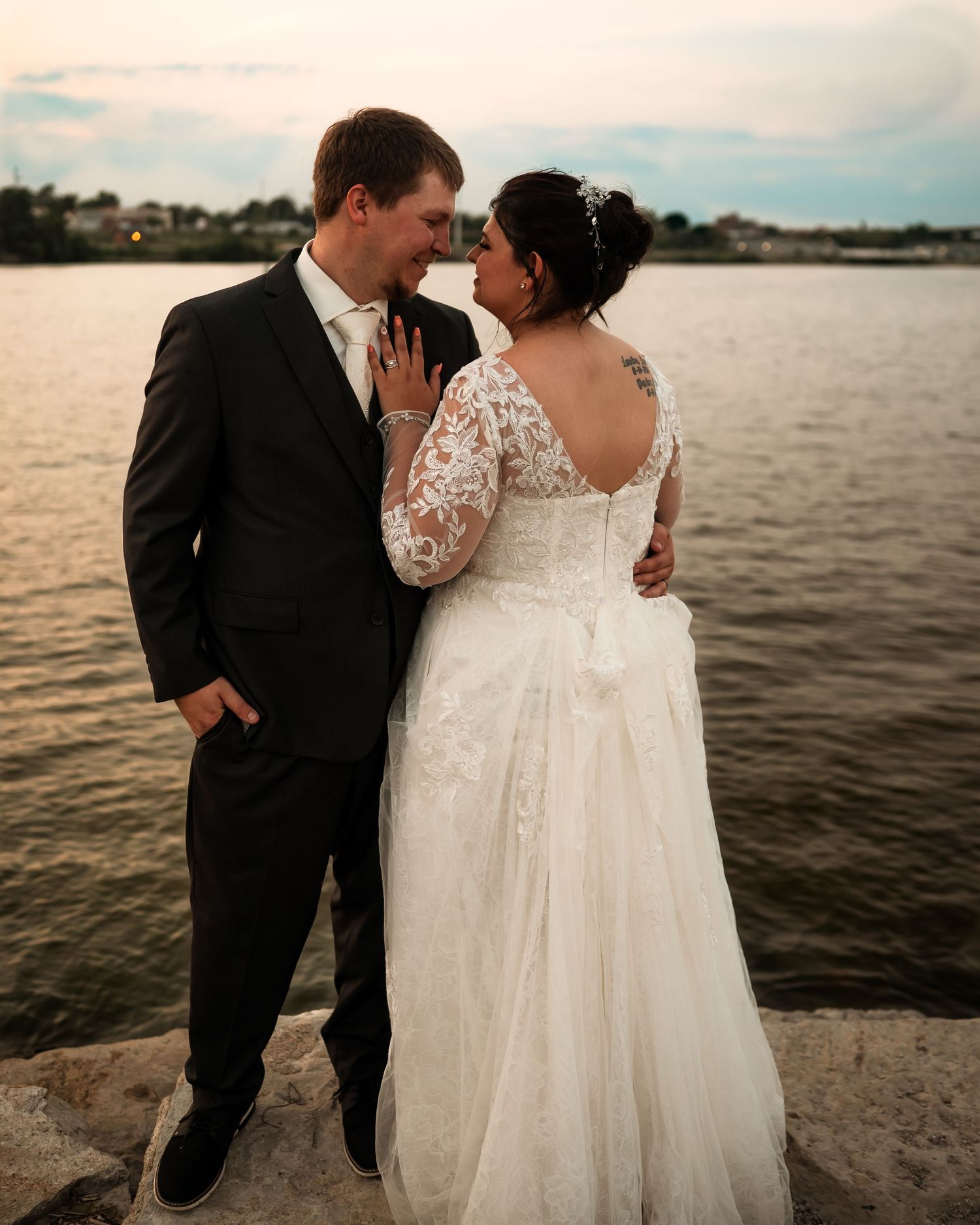 A romantic wedding couple embraces by the waterfront at sunset, with the bride wearing a lace gown.