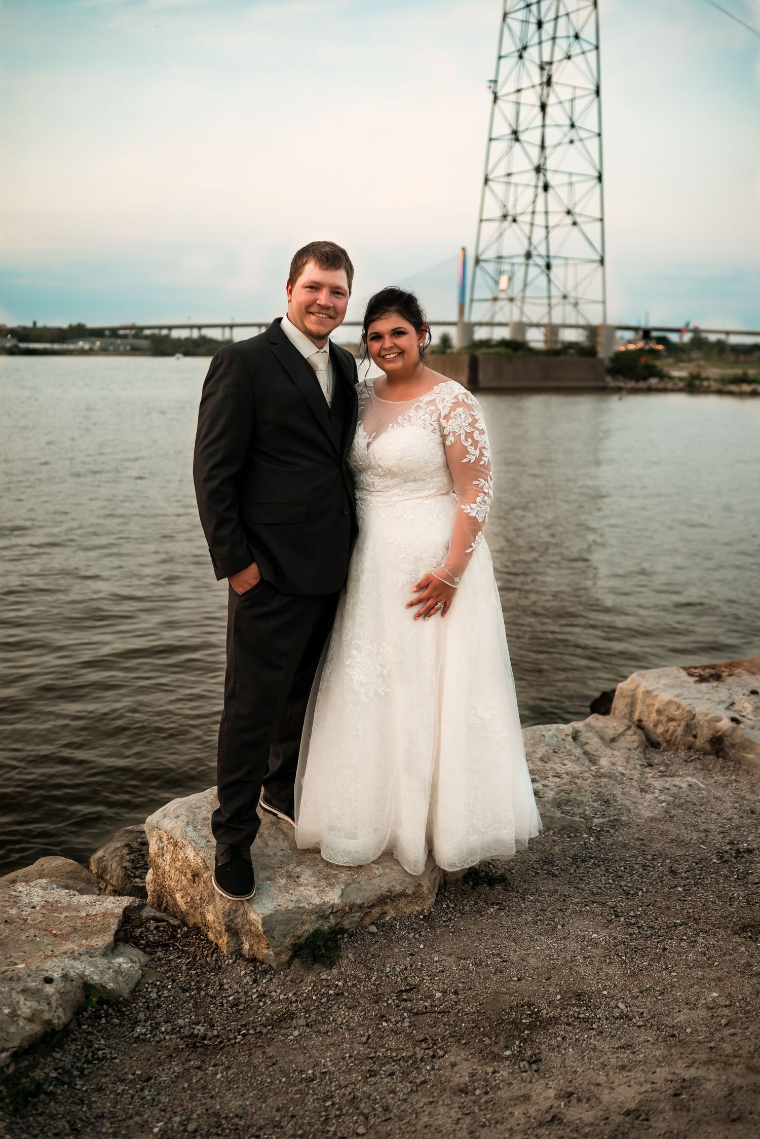 Wedding couple poses near industrial towers by the waterfront in their formal attire.