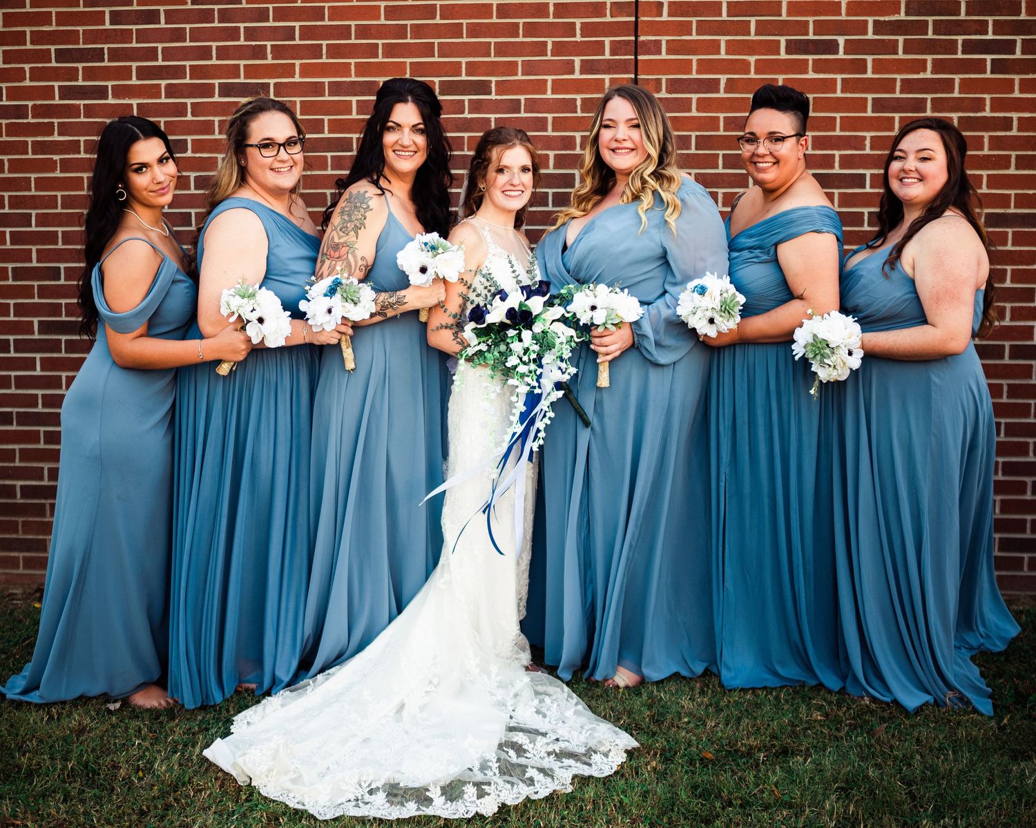 Wedding party in coordinated blue dresses and white bouquets stand together outdoors.