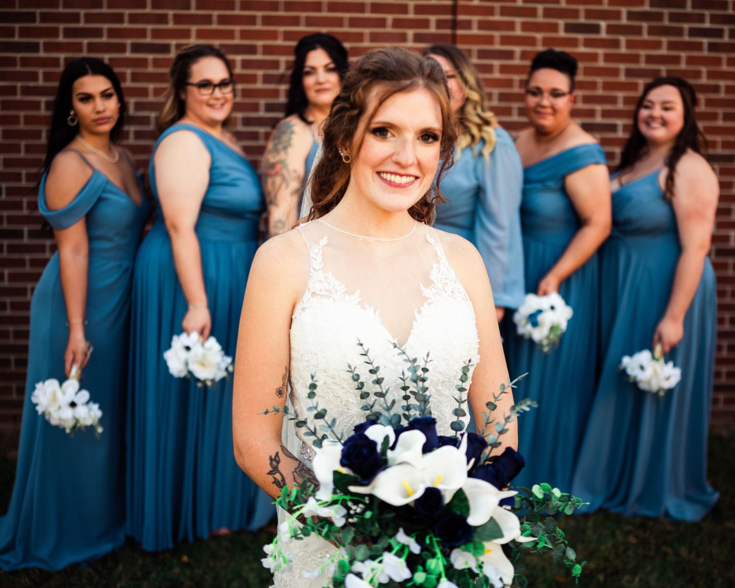 Bridesmaids in flowing blue dresses pose with white bouquets against a brick wall.